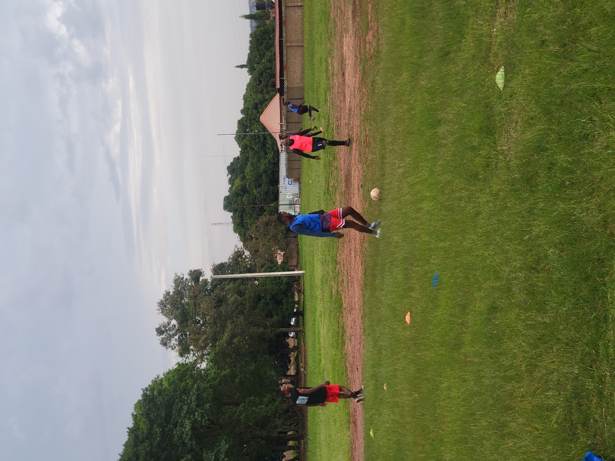 This image depicts an outdoor scene on a grassy field, likely a sports ground, in Jos, Nigeria. The main subjects are several individuals, appearing to be young men, engaged in what looks like a soccer training session. They are dressed in athletic wear, including shorts, t-shirts, and vests in various colors like blue, red, and pink. One individual in the foreground is lying on the grass, while others are standing or in motion, suggesting drills or practice. A soccer ball is visible on the field, reinforcing the sporting context.
The setting includes a fence in the background, possibly surrounding the field, and a building with a red roof. Lush green trees form a backdrop, and in the distance, there are indications of urban structures, including what appears to be a telecommunications tower. The weather seems overcast, with a pale, cloudy sky dominating the upper portion of the image, suggesting it might be daytime, possibly late morning or afternoon. The ground is a mix of green grass and patches of reddish-brown soil, perhaps indicating areas of heavy use or wear on the field. There are also several small cones scattered across the field, commonly used as markers in sports training.
No discernible text is visible in the image. The overall mood appears to be one of focused activity and athletic exertion.