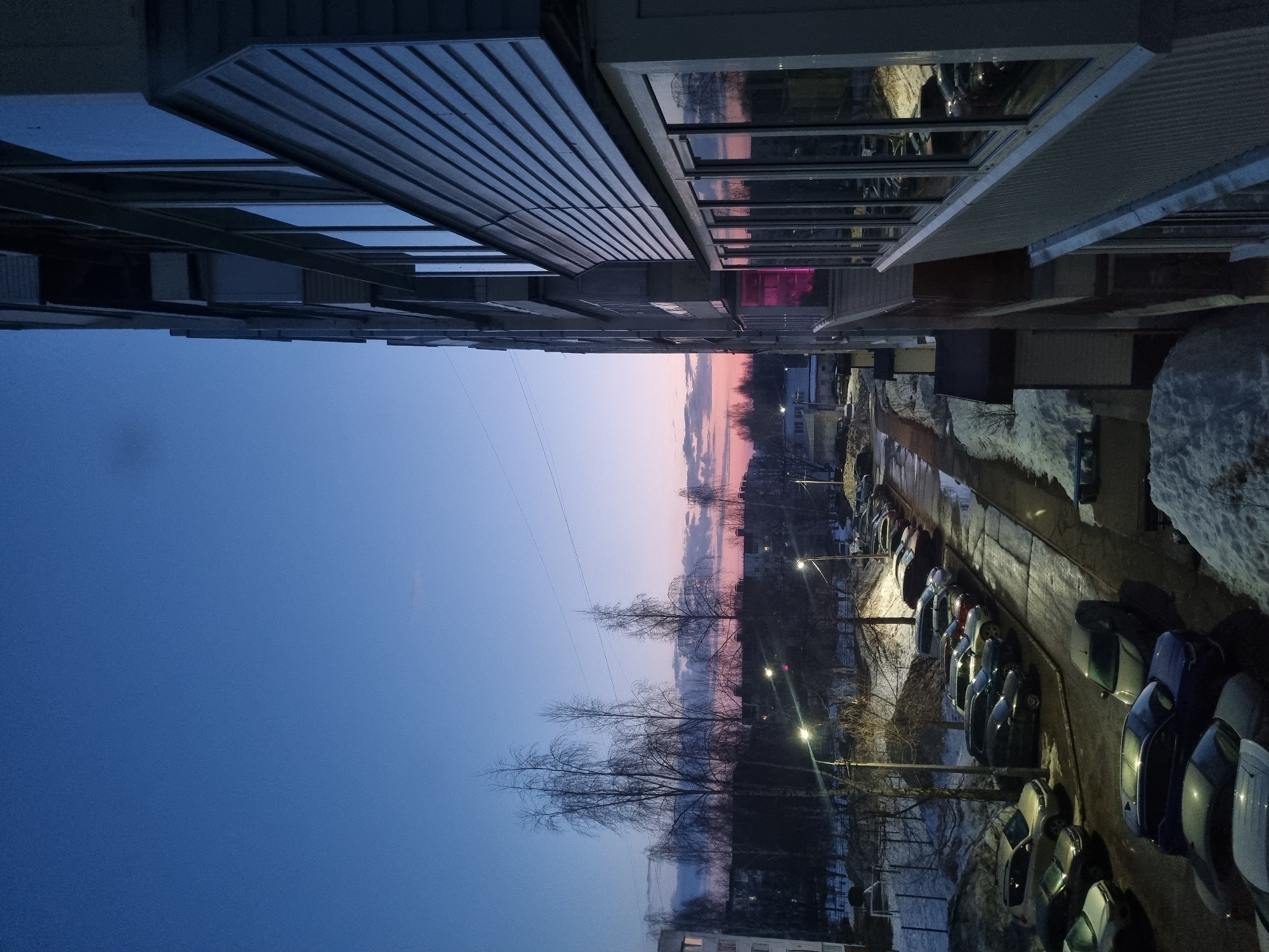 View looking out from between two buildings in Chaikovsky, Russia at dusk. The sky transitions from blue to pink on the horizon. Bare trees are in the center. Streetlights line the street next to a row of parked cars. Patches of snow are visible. The buildings on either side have rows of windows, some reflecting light.
