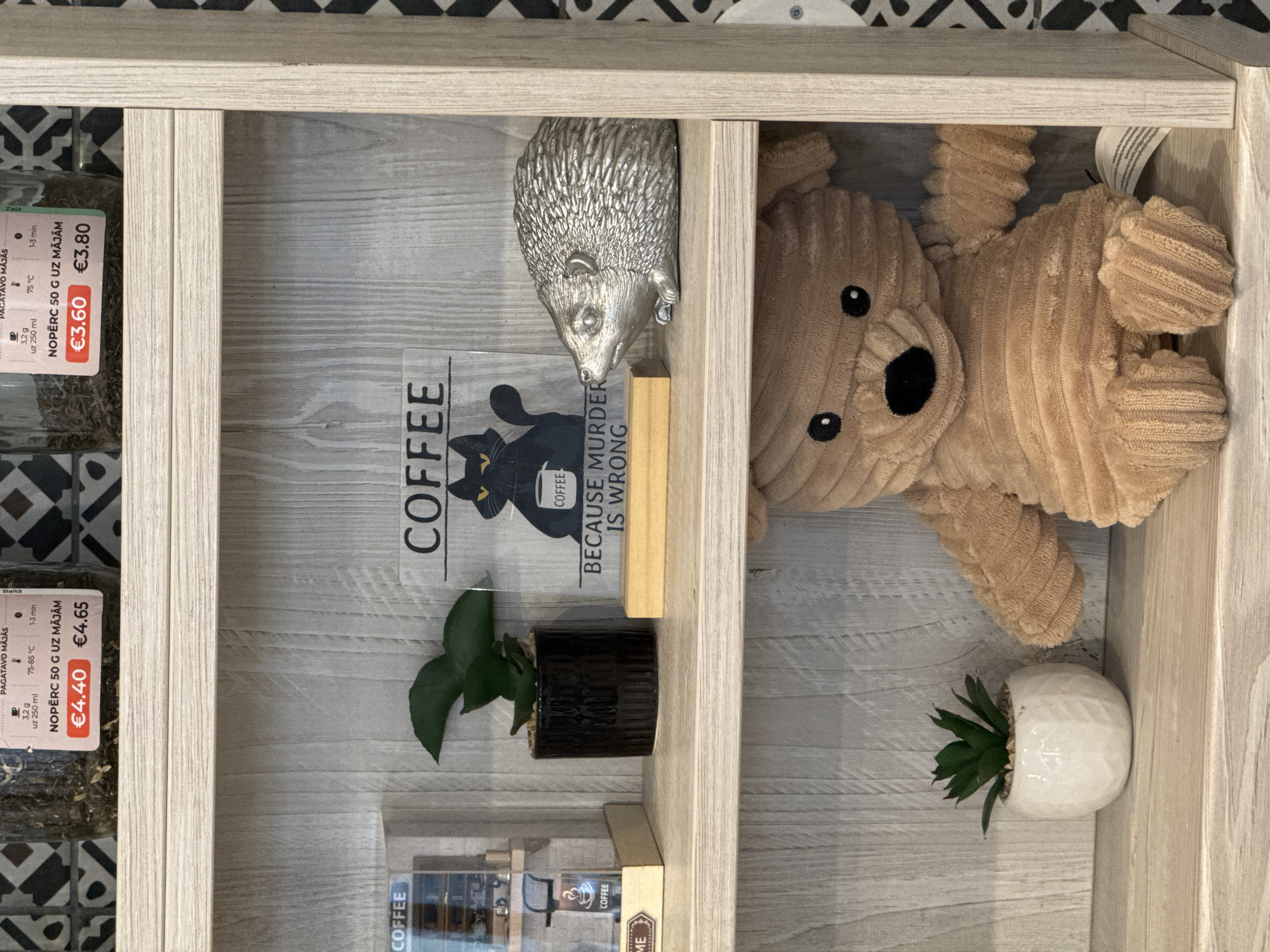 The image displays a multi-compartment wooden shelving unit, likely part of a retail display in a shop or cafe in Riga, Latvia. The background behind the shelves features a black and white patterned tile or wallpaper.
On the top shelf, the left compartment holds two clear jars filled with dried herbs or tea. Their labels, partially in Latvian, read "PAGATAVO MĀJĀS" (Prepare at Home) and "NOPĒRC 50 G UZ MĀJĀM" (Buy 50g for home), along with preparation details like "3,2 g uz 250 ml", "75 °C", "1-3 min", and prices €3.60 and €3.80. The right compartment on the top shelf contains a silver hedgehog figurine, partially tucked under the shelf above, and a plush, light brown teddy bear with a corduroy texture, resting on its side. The bear has black button eyes and a black nose. A small, green succulent plant in a white, faceted pot is also visible here.
On the bottom shelf, the left compartment features another jar of tea/herbs with similar Latvian labels, including "75-85 °C" and "1-3 min" for preparation, and prices of €4.40 and €4.65. Next to this, a prominent framed sign displays "COFFEE" at the top, a black cat looking into a coffee cup, and the phrase "BECAUSE MURDER IS WRONG" below it. A small plant in a black ridged pot sits in front of the sign. The right side of the bottom shelf, partially visible, contains more items, including labels with the word "COFFEE" and a small sign reading "ME". The overall impression is a cozy and curated retail display.