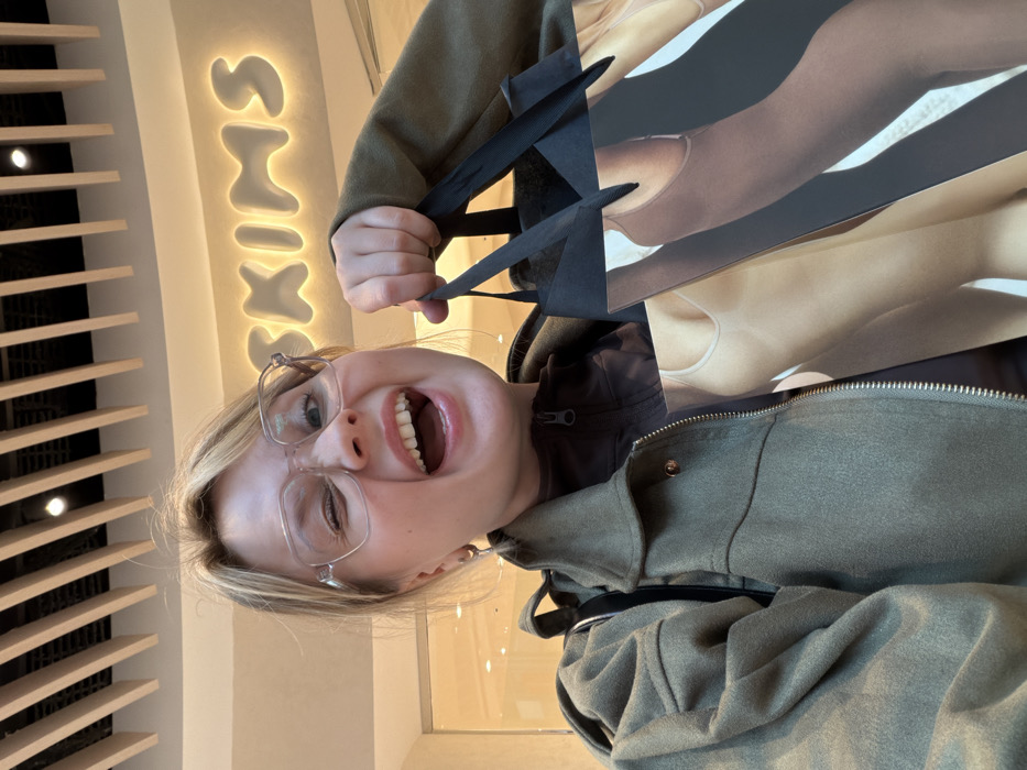 A close-up selfie depicts a young Caucasian female, mid-torso up, in an indoor retail environment. She has blonde hair, wears clear-framed glasses, an olive-green jacket over a dark brown zipped top, and a silver hoop earring in her left ear. Her mouth is wide open, teeth visible, and her left eye is winking, conveying an excited expression. She holds a black-handled shopping bag in her right hand, printed with an artistic depiction of torsos wearing neutral-toned, form-fitting garments, characteristic of the SKIMS brand. The background features a brightly illuminated "SKIMS" sign with bubble lettering. The ceiling is composed of parallel light-colored slats with integrated lighting. This scene is located within a shopping mall in Tel-Aviv, Israel, consistent with the SKIMS store in the Fashion Mall.