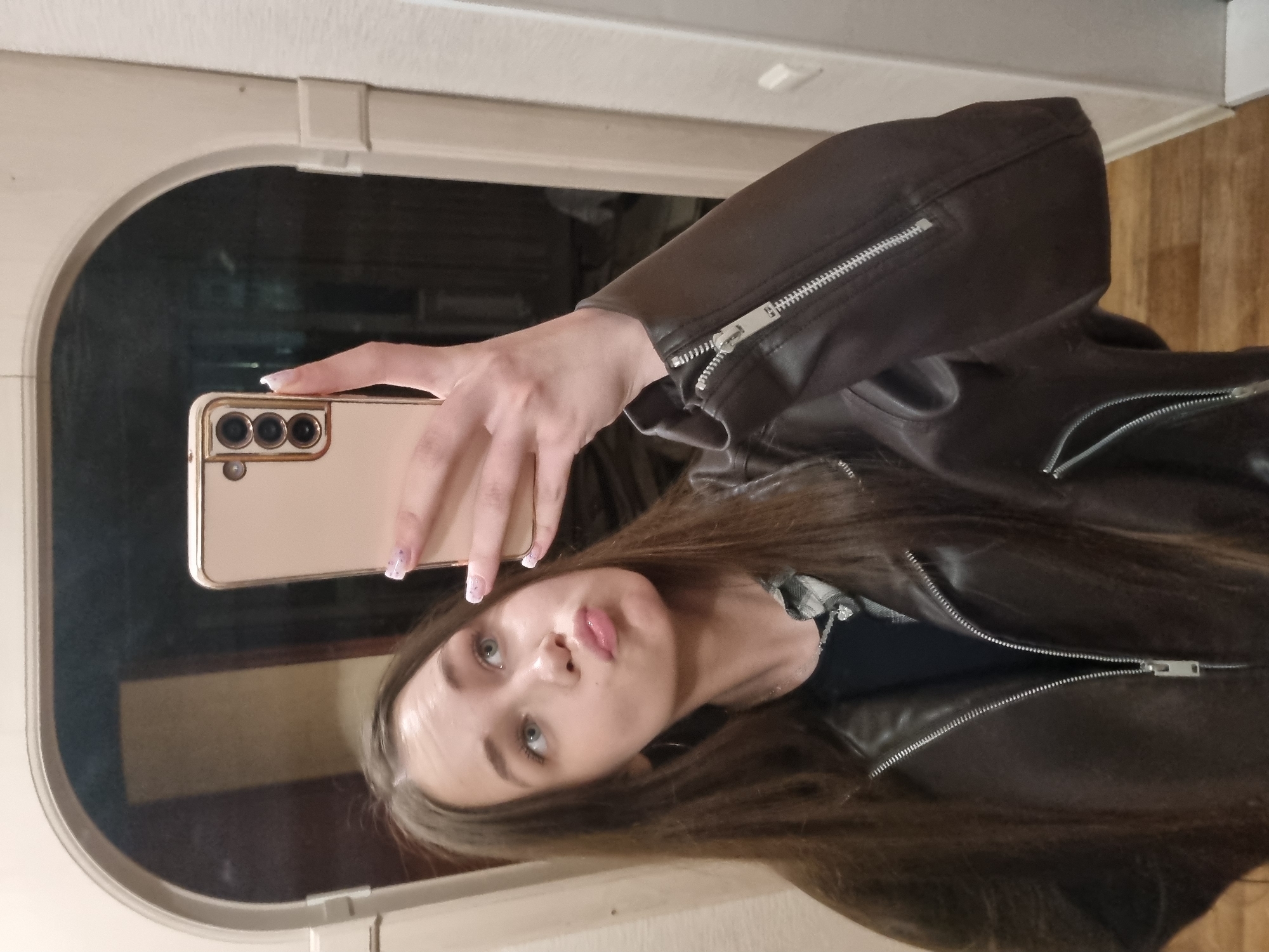 A rotated, eye-level mirror selfie features a young woman indoors. She has long, straight dark brown hair and light-colored eyes, with her lips pursed in a playful pouting expression. She is wearing a dark brown leather jacket with prominent silver zippers on the sleeves and body. A plaid shirt is visible beneath the jacket, worn over a dark top.
The woman holds a gold or beige-colored Samsung smartphone in her right hand, with its three-lens camera module clearly visible. Her nails are manicured with light-colored polish adorned with small, decorative specks.
The background, viewed through the mirror's reflection, includes a distinctive light-colored, possibly off-white or beige, arched frame at the top, likely part of a doorway or the mirror itself. A darker, indistinct area is reflected behind her, and a light-colored wooden floor is reflected on the right side of the image. The overall setting is casual and indoors. No visible text is present.