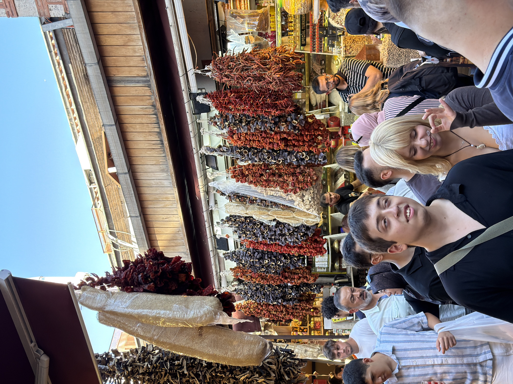 This image captures a vibrant scene at the Spice Bazaar in Istanbul, Turkey, a bustling marketplace known for its aromatic spices and dried goods. The foreground is filled with a diverse crowd of people, their faces showing various expressions of engagement and curiosity.
In the center, a young woman with blonde hair smiles and makes a gesture with her hand, possibly indicating approval or a small item she holds. To her left, a young man in a striped shirt carries a white plastic bag, his gaze directed forward. Behind them, an older man with a grey beard looks towards the right, his expression unreadable.
The background is dominated by the colorful displays of dried goods hanging from the ceiling. Bunches of red chili peppers, dried eggplants, and other produce create a rich tapestry of textures and colors. These are characteristic of the Spice Bazaar, where vendors sell a wide variety of spices, dried fruits, nuts, and Turkish delight. The stalls are adorned with shelves stocked with jars and bags of ingredients, hinting at the culinary delights available.
The architecture of the bazaar is visible, with wooden beams and awnings overhead, typical of traditional covered markets. The lighting suggests it is daytime, with sunlight filtering through the market's openings, illuminating the scene and casting shadows. The overall atmosphere is one of lively commerce and cultural immersion, with the sensory experience of the market emphasized by the visual abundance of goods and the presence of shoppers.