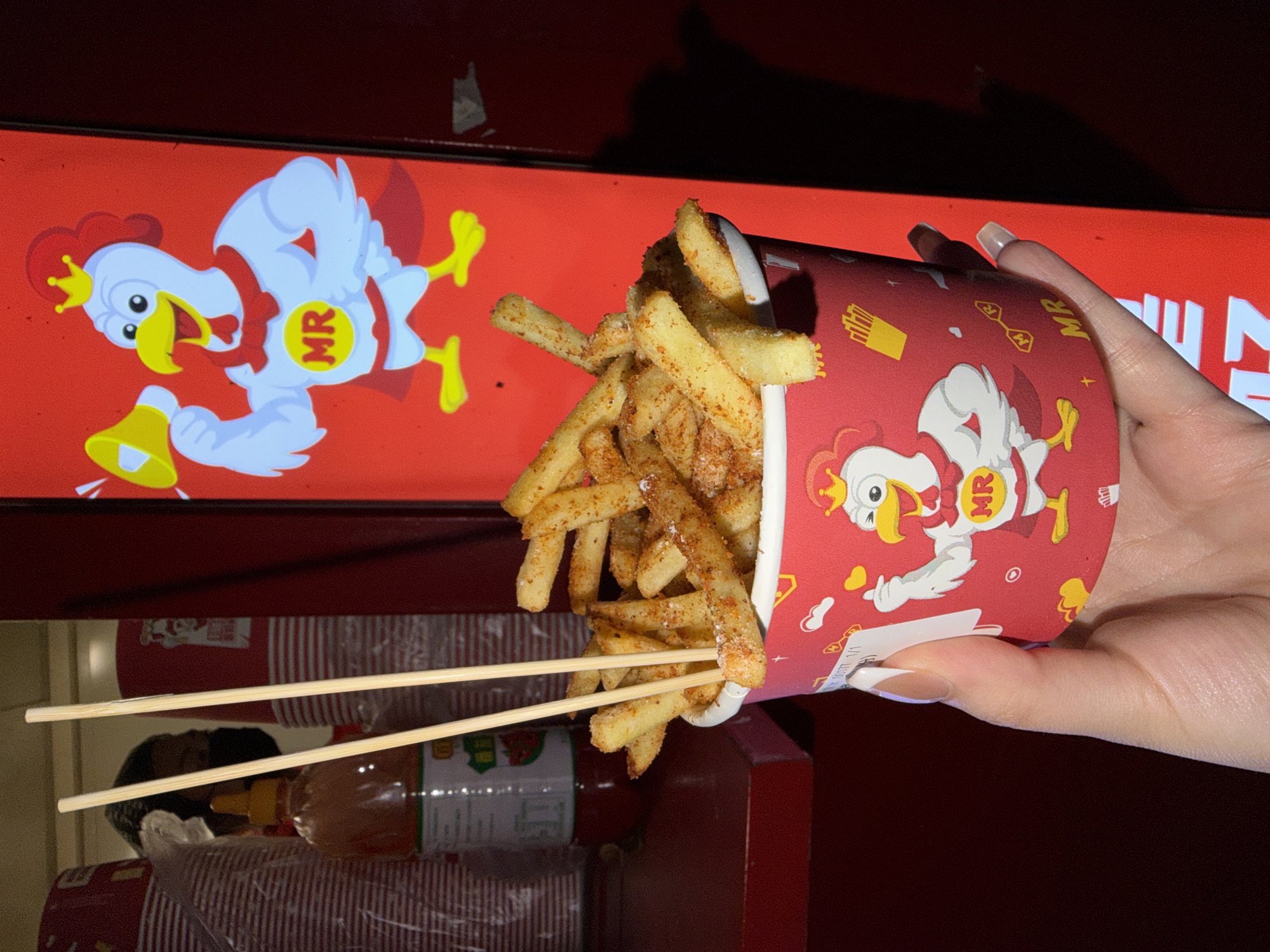 A close-up, indoor shot shows a hand holding a red and white paper cup filled with French fries, with chopsticks protruding from the fries. The cup features a cartoon chicken wearing a crown and a cape with the letters "MR". In the background, a red signboard displays a similar chicken cartoon and Chinese characters. Below the cup, a shelf contains bottles of sauce and other packaging. The scene appears to be in Chongqing High-tech Zone, China.