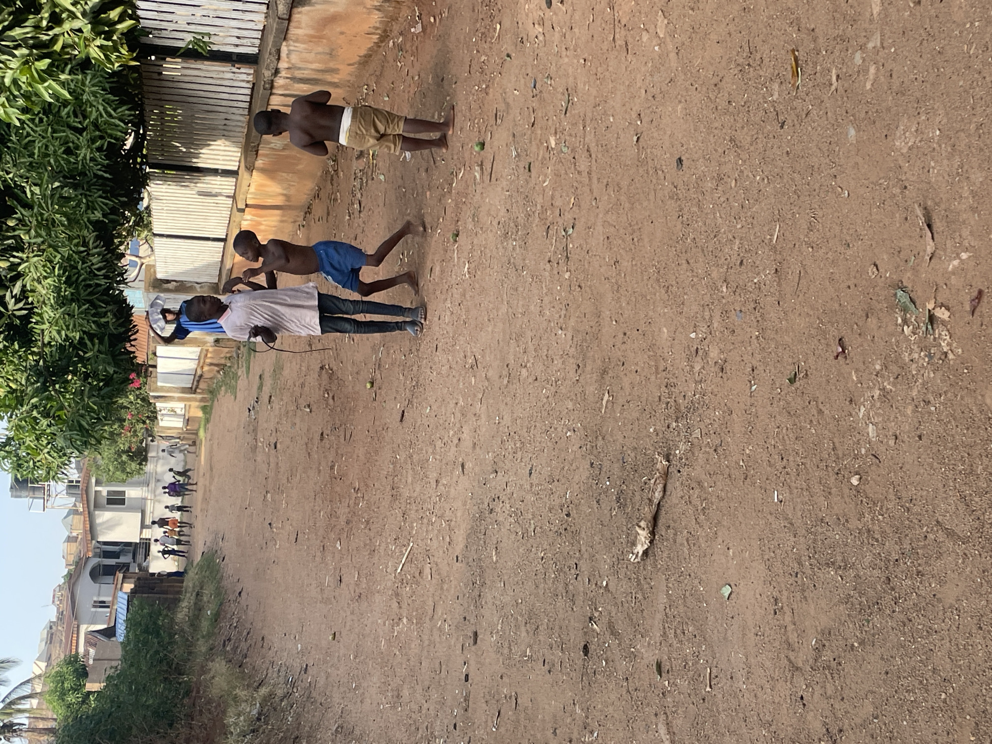 Three children are in a bare, sunlit dirt lane in Jos, Nigeria. One child, wearing a white shirt and dark pants, is in the foreground. A second child is wearing a blue shirt and is positioned behind the first child. A third child, with no shirt and tan shorts, is further ahead. The children appear to be playing. A fence and building are on the left side of the lane and trees/shrubs are on the left and right side of the lane. Buildings can be seen in the distance on the left side of the lane.