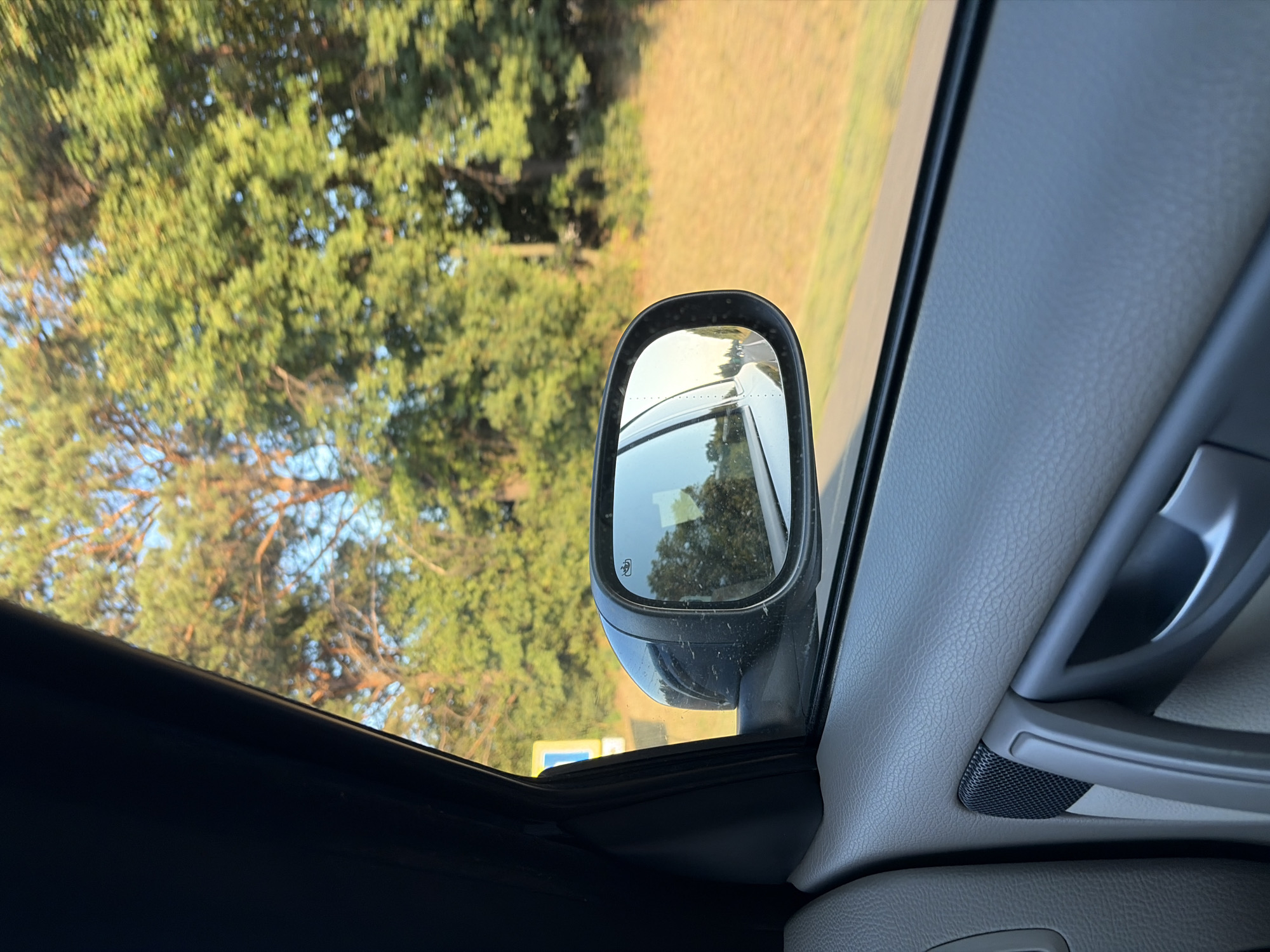 The image, taken from inside a vehicle, shows a side mirror reflecting trees and a road. The interior of the car, including a door handle and speaker grille, is visible in the foreground. Outside the car, the trees are lush and green, and the sky is a clear blue. A yellow road sign with a blue rectangle is visible in the distance on the left side of the image. The scene appears to be a roadside landscape.