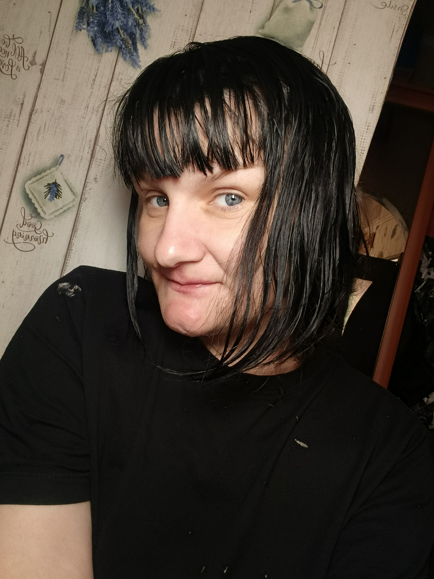 A close-up shot of a woman with wet, short black hair and bangs. She has fair skin and blue eyes. She is wearing a black t-shirt with what appears to be paint or debris on the shoulder and front.
The background includes a wooden-patterned wall with floral decorations. Part of what appears to be furniture is visible on the right side of the frame.
There is no information to determine if this image was taken in Фоки, Russia.
