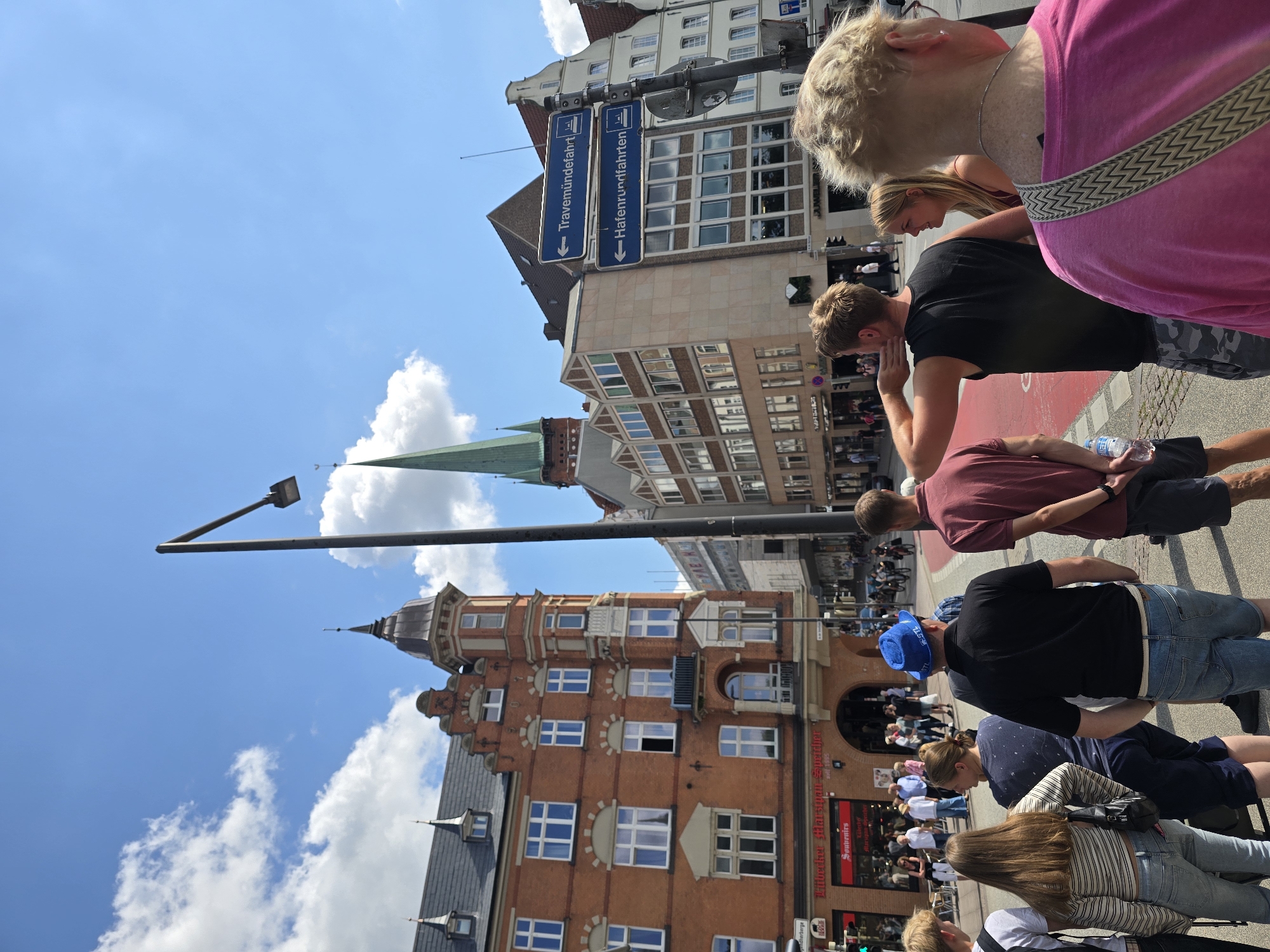 The image depicts a street scene in Lübeck, Germany, on a sunny day with scattered clouds. In the foreground, a group of people are gathered on a street. To the left, an older brick building with multiple windows and ornate architectural details is visible. Further back, a green-spired church tower dominates the skyline. Several other buildings with various architectural styles line the street. Traffic signs, one indicating "Travemündefahrt" and another "Hafenrundfahrten," are mounted on a pole. Pedestrians and some cyclists are visible further down the street, suggesting a public area. A person in the foreground is holding a bottle of water. The overall atmosphere appears to be a bustling urban environment.