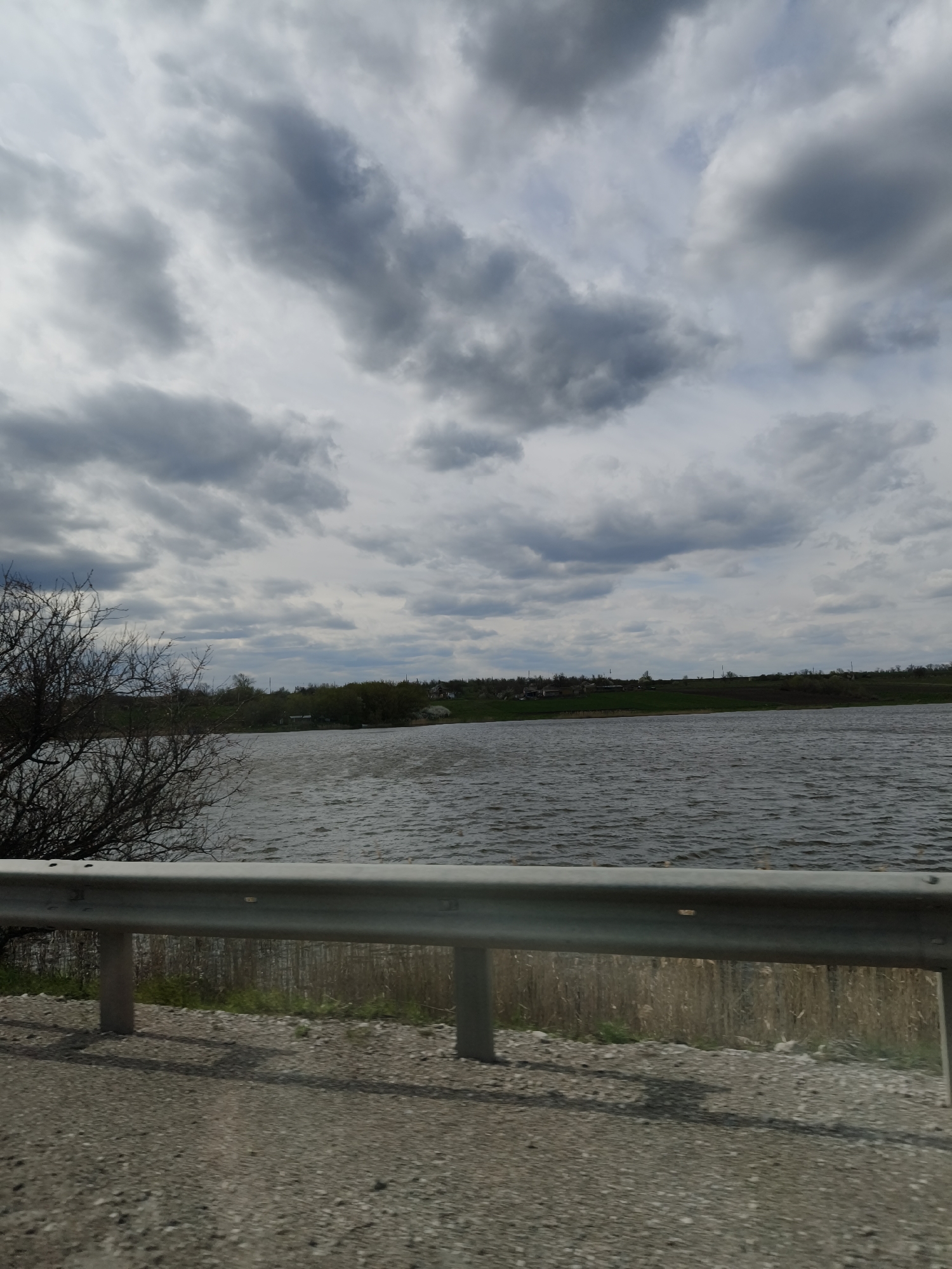 This media file captures a wide landscape scene featuring a body of water, land, and an overcast sky, likely taken from a roadside perspective in Sofiivka, Ukraine.
In the foreground, a gray metal guardrail stretches horizontally across the lower part of the frame, indicating the edge of a road or viewing area. Below the guardrail, there is a visible gravel or dirt road shoulder. Behind the guardrail, some dry, light-colored reeds or tall grasses can be seen near the water's edge. To the left, a portion of a tree with bare, dark branches extends into the frame.
The midground is dominated by a large body of water, possibly a lake or wide river. The water's surface is textured with numerous ripples and small waves, suggesting windy conditions.
In the background, on the opposite bank of the water, a relatively flat expanse of land is visible. This land features patches of green grass and scattered trees, indicating a natural or agricultural landscape. Several small, distant buildings, likely houses, are dotted along the far shore, suggesting a rural settlement or village.
The sky is heavily overcast with dense, grey clouds that fill the upper portion of the image. While the clouds are predominantly dark grey, some lighter grey patches suggest varying thickness or breaks in the cloud cover. There is no direct sunlight visible, indicating a cloudy daytime.
No specific human or animal activity is depicted. There is no legible text visible in the image. The overall impression is one of a quiet, rural landscape on an overcast day.