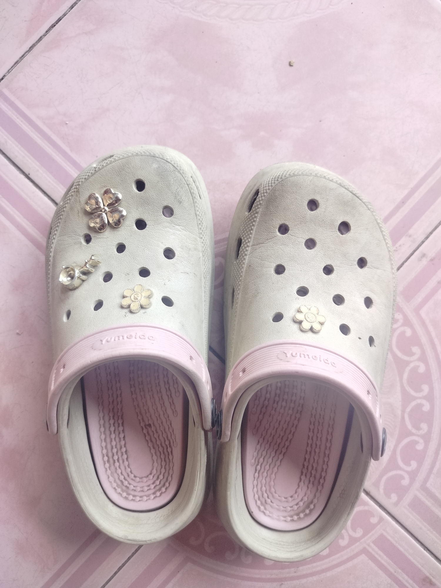 A pair of light-colored clogs with pink accents and decorative charms sit on a tiled floor. The clogs are a size large and appear to be for a child, judging by the small size and cute charms. The left clog has a silver four-leaf clover charm and a silver snail charm, while the right clog has a white flower charm. The floor is tiled in a light pink and white pattern, with some scuff marks visible. The overall scene suggests a domestic setting, possibly a child's room or play area in a home in Kumbangindah, Indonesia.