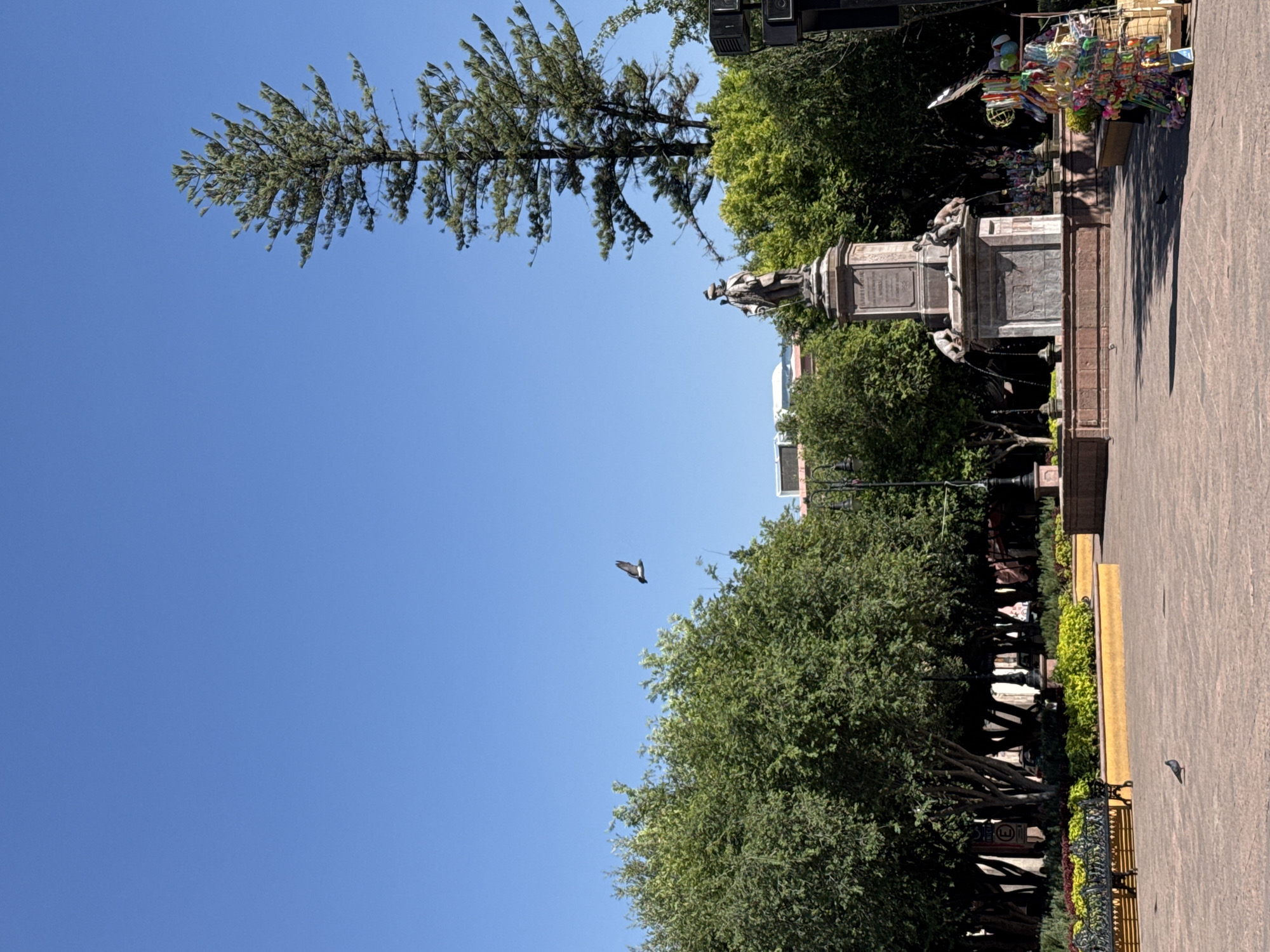 The image shows a sunny day in the city of Querétaro, Mexico. The camera looks up to capture a statue or monument, surrounded by trees and a clear blue sky. A bird is flying in the middle-ground. The statue is mounted on a stone base, featuring decorative elements and possibly water features. Vendors with colorful merchandise occupy the edge of the plaza area near the monument. Paved ground covers the area in front of the statue, where a bench is positioned along a landscaped border.