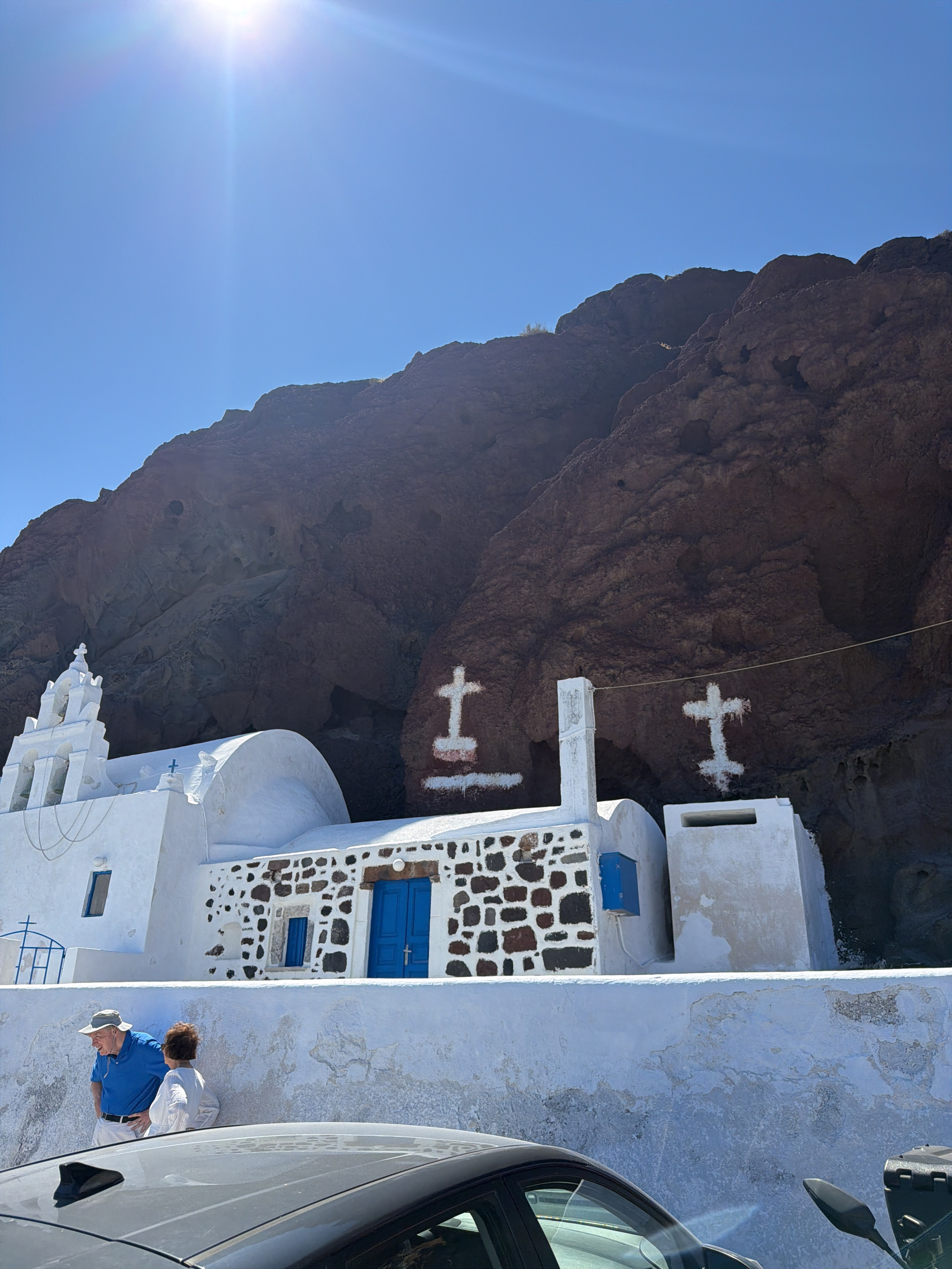 A bright sun shines on a whitewashed church built into the side of a dark, rocky cliff in Thira Municipal Unit, Greece. The church has a bell tower with three bells and a dome. Two figures stand on a ledge in front of the church, looking at something to their left. A dark car is parked in the foreground. The cliff face is marked with two white crosses.