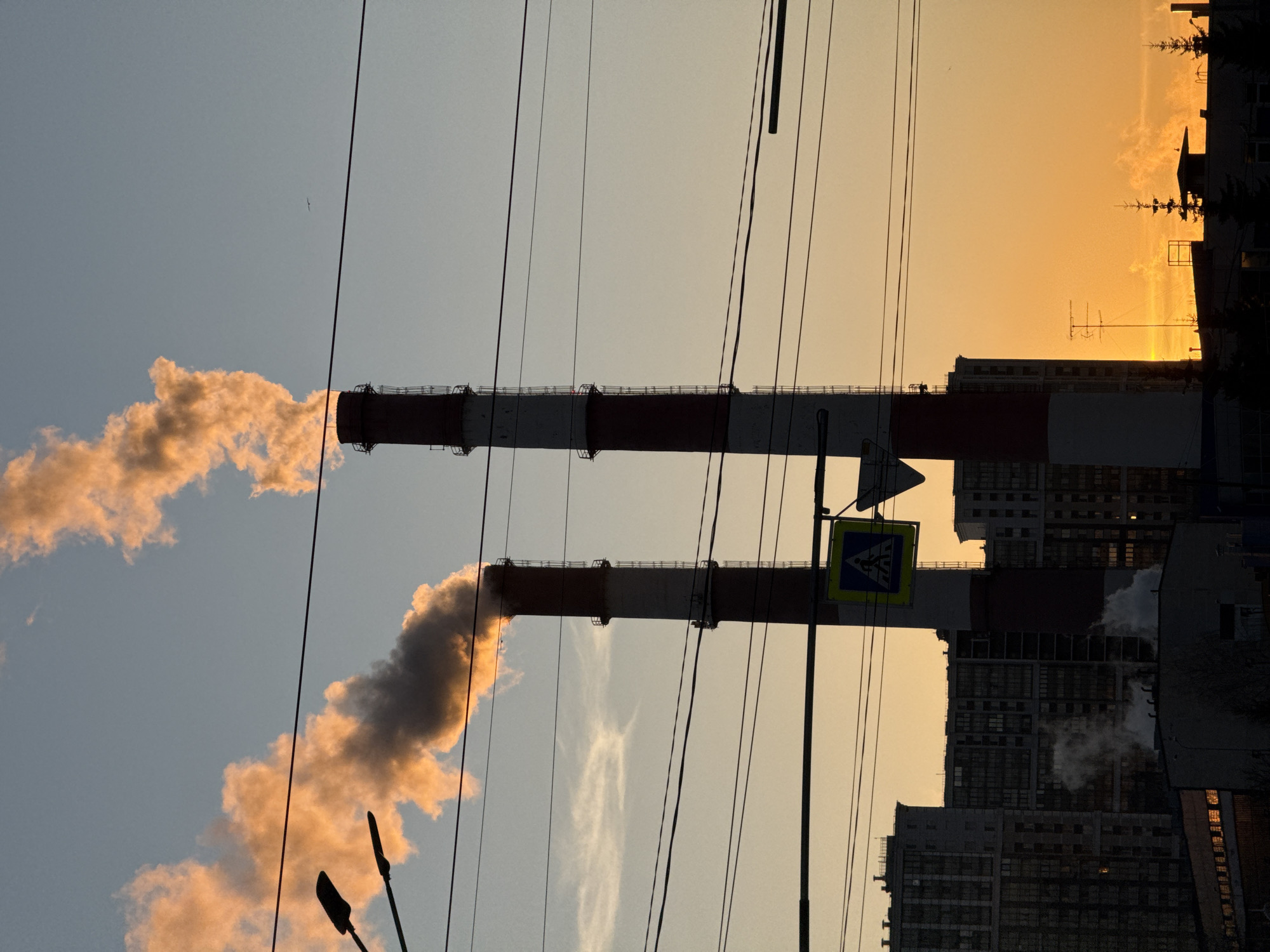 The image shows two tall industrial chimneys emitting smoke or steam, located in an urban area, Saint Petersburg, Russia. The chimneys are painted with alternating red and white stripes. Power lines crisscross the scene. In the background are buildings and a partly-obscured sunset. A pedestrian crossing sign is visible.
