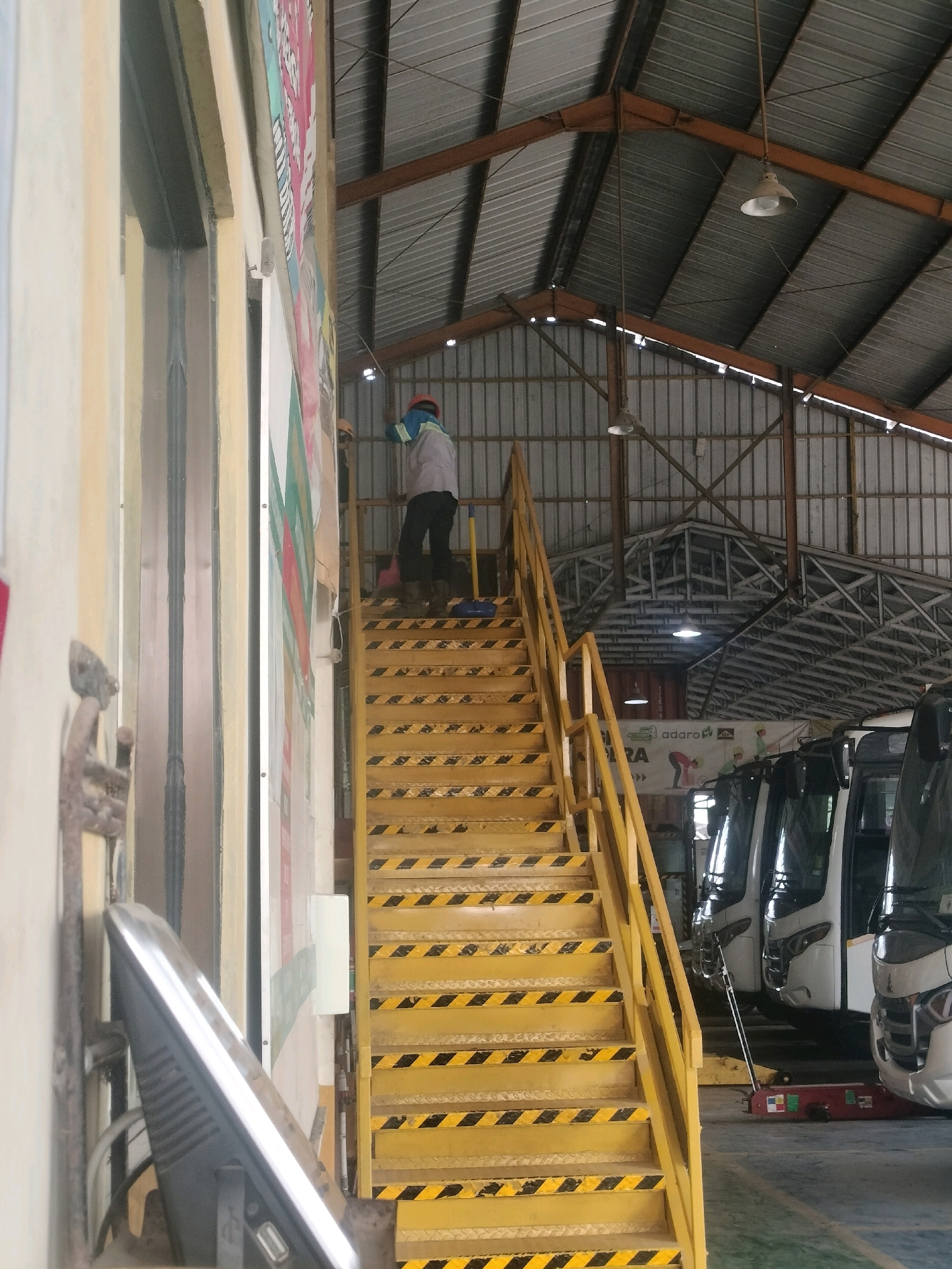 In Maburai, Indonesia, a person wearing a hard hat and safety vest stands on a yellow staircase with black hazard stripes. They are looking down as they ascend the stairs, which are part of an industrial building with a corrugated metal roof and exposed beams. The staircase leads to an upper level, out of frame. In the background, several white buses are parked in a hangar-like structure. The environment appears to be a maintenance or service area for the buses.