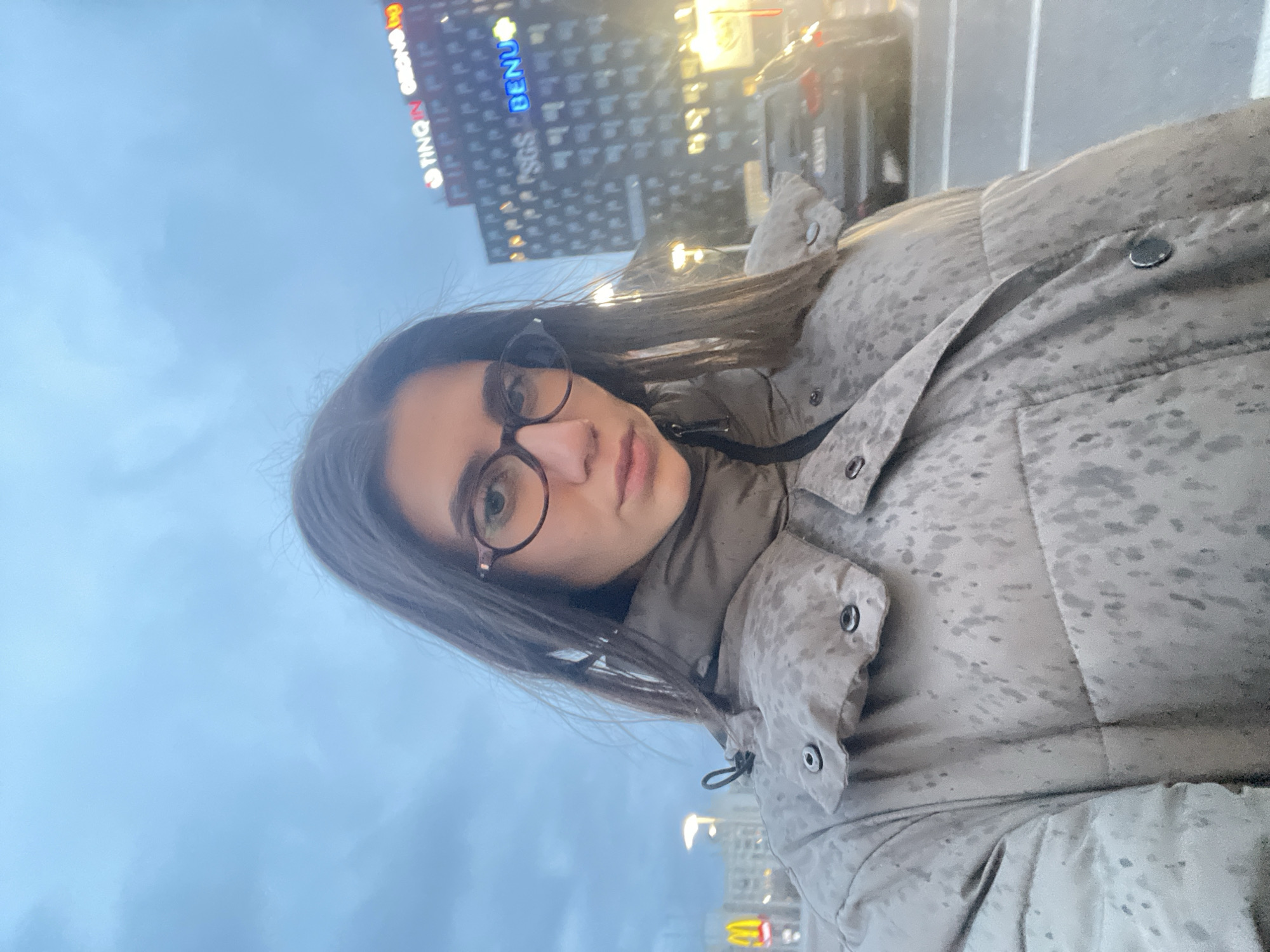 A selfie shows a woman with light skin, dark hair, and glasses. She is wearing a camouflage-patterned jacket with snap closures. Behind her is a blurred cityscape under a cloudy sky. A building with neon signs reading "TING-IN GEORGE" and "BENU" is visible. The scene is identified as Sofia, Bulgaria.