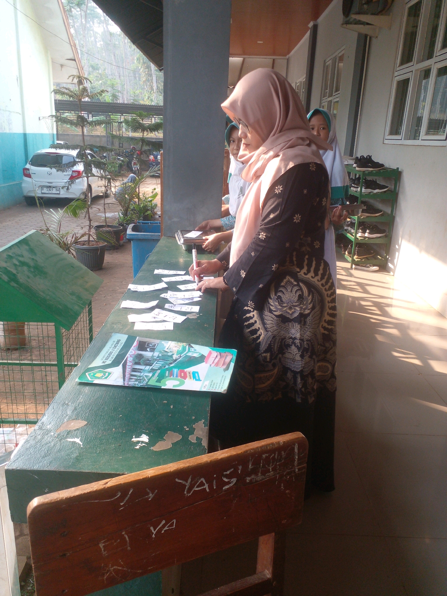 In front of a building with a covered walkway, possibly at a school in Banyuputih, Indonesia, a woman wearing a light pink hijab and a dark dress with gold patterns is standing at a green table. She is holding a red marker and appears to be writing on small pieces of paper scattered on the table. Next to her, a younger woman in a white school uniform and light blue hijab is looking towards the camera. Another person, mostly obscured, is also visible behind the woman in the pink hijab. On the table, there is a magazine or booklet with the title "KEMENTERIAN AGAMA KABUPATEN BATANG". To the left of the table, there is a green cage-like structure and several potted plants. In the background, a white car with license plate "H 3329 BW" is parked. Further back, there are trees and other buildings. Sunlight is casting shadows on the tiled floor. A wooden chair with graffiti is positioned in the foreground.