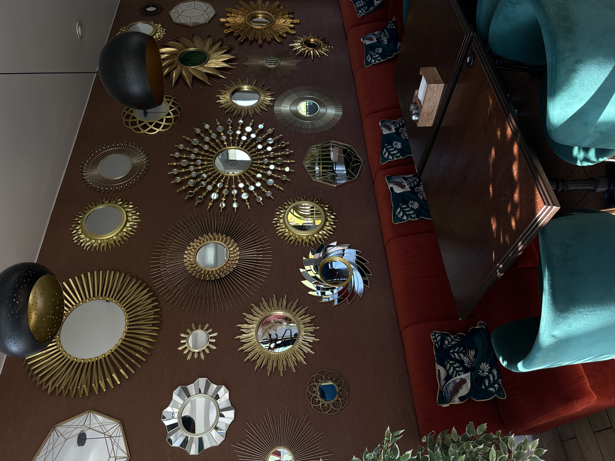 The image depicts the interior of what appears to be a restaurant or café. The scene is oriented from an elevated perspective, looking down at a wall, furniture, and decor. The wall is painted in a dark brown color and is adorned with a collection of ornate, mostly circular mirrors with golden or silver frames of varying designs. On the left, there are two large, black and gold lighting fixtures. To the right of the wall is a red cushioned banquette seating area with patterned throw pillows. A dark wooden tabletop is positioned adjacent to the seating, indicating a dining area. The chairs around the table are upholstered in a vibrant teal fabric. Plant life is visible in the foreground, adding a touch of greenery to the setting. The location is likely Chaikovsky, Russia based on the prompt instructions, though there are no inherent clues to confirm this detail.