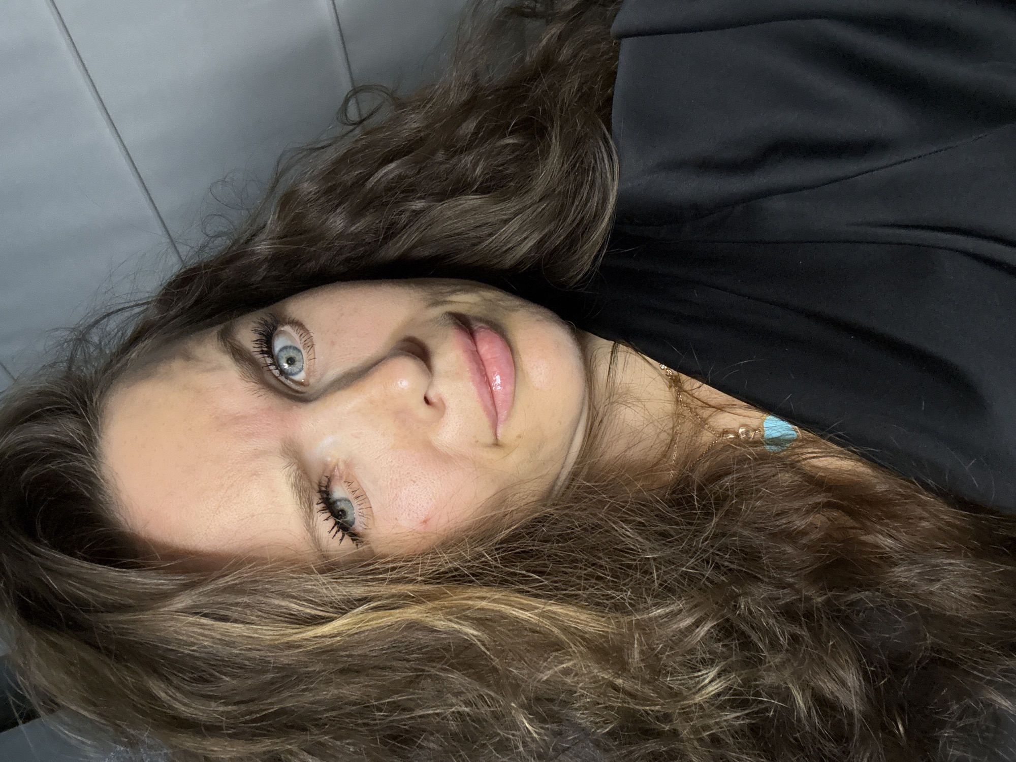 A close-up photograph, rotated counter-clockwise, depicts a person with light skin and blue eyes, presumably female, with shoulder-length, wavy brown hair. The person's face is prominently featured, with a slight smile. The background is not clearly defined, but includes a dark solid fabric, likely clothing. A gold chain necklace with a light blue stone is visible near the neck. The location cannot be determined from this image.