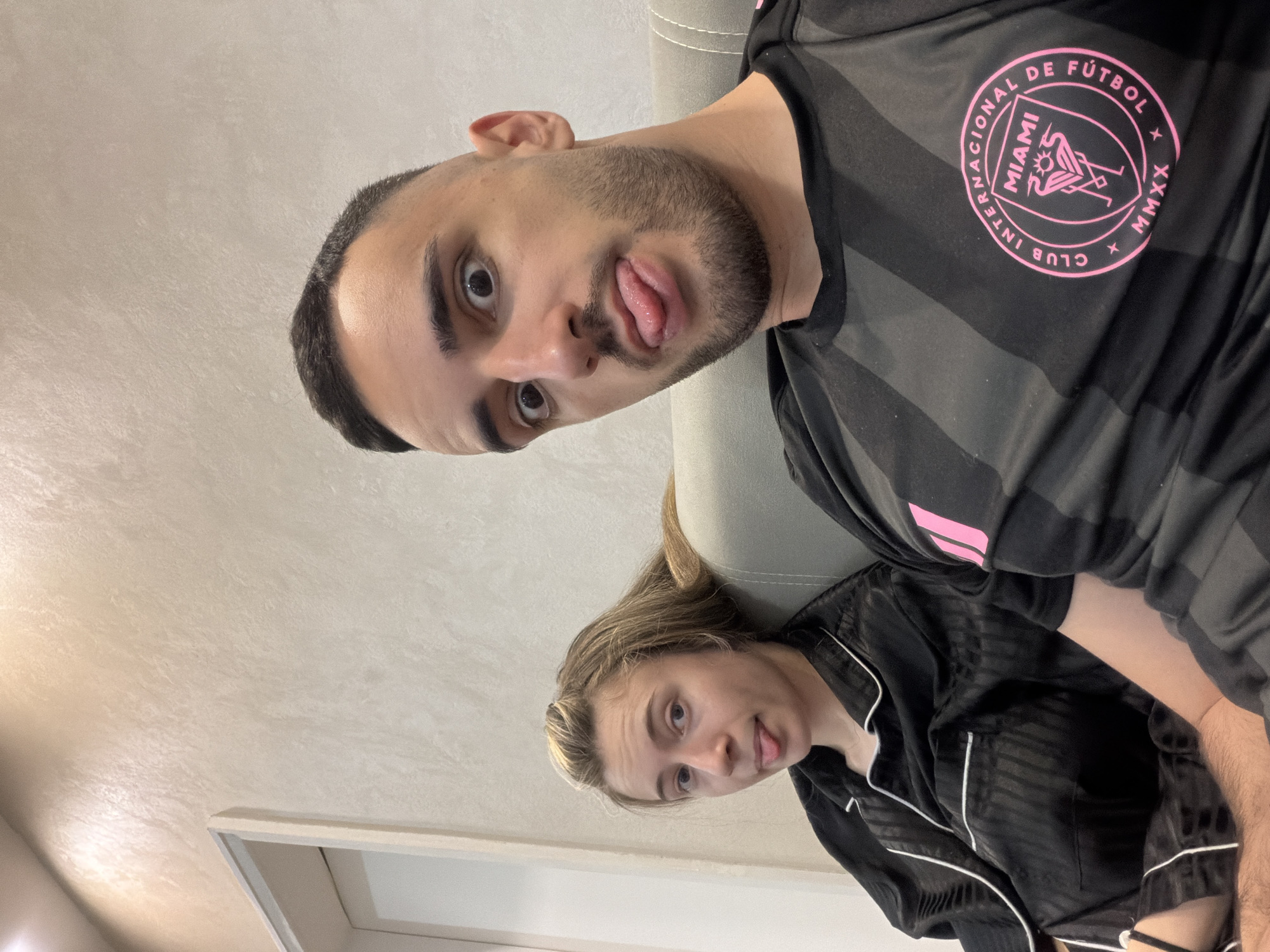 A photo shows a man and a woman, both sticking their tongues out, likely in a selfie taken indoors. The man is wearing a black soccer jersey with the Inter Miami logo. The woman is wearing a black pajama top with white piping. The location cannot be determined as Plovdiv, Bulgaria.