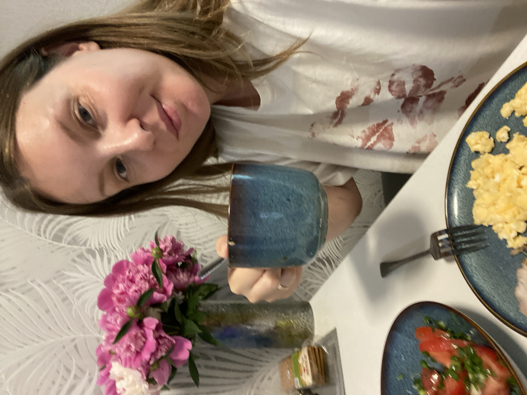 The image shows a woman in Noviy, Russia, sitting at a table with a meal and a bouquet of pink peonies. She is holding a blue mug and looking at the camera. The table has two plates of food, one with scrambled eggs and another with tomatoes and green onions, and a fork. The woman is wearing a white t-shirt with red lipstick marks. The background shows a wall with a palm leaf pattern.