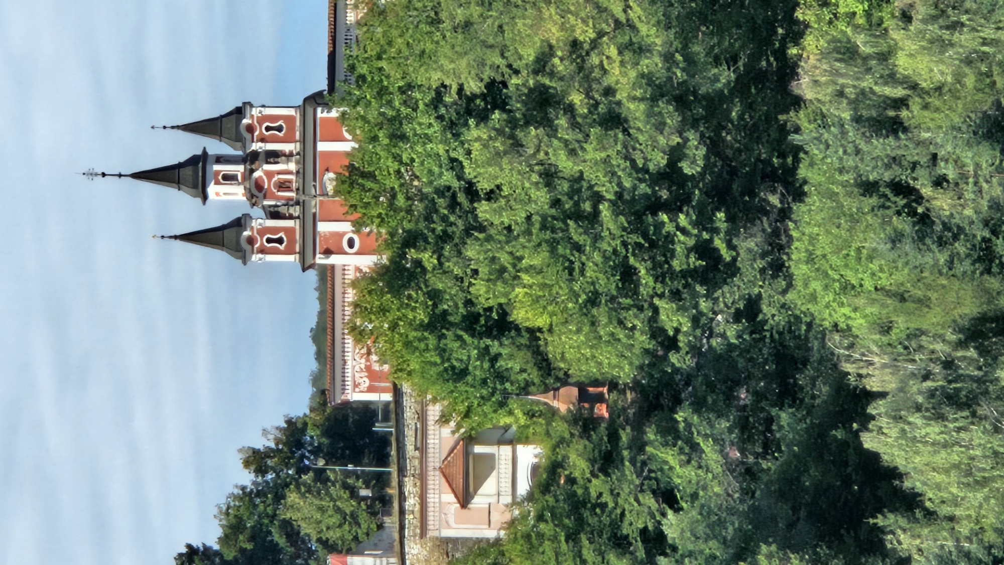 The image shows the Church of St. Nicholas in Prešov, Slovakia. The church is a historic landmark in the city and is known for its distinctive red and white architecture and its three spires. The church is surrounded by lush green trees, and the sky is a clear blue with some scattered clouds. The image captures the church from a distance, giving a sense of its grandeur and its setting within the natural landscape.