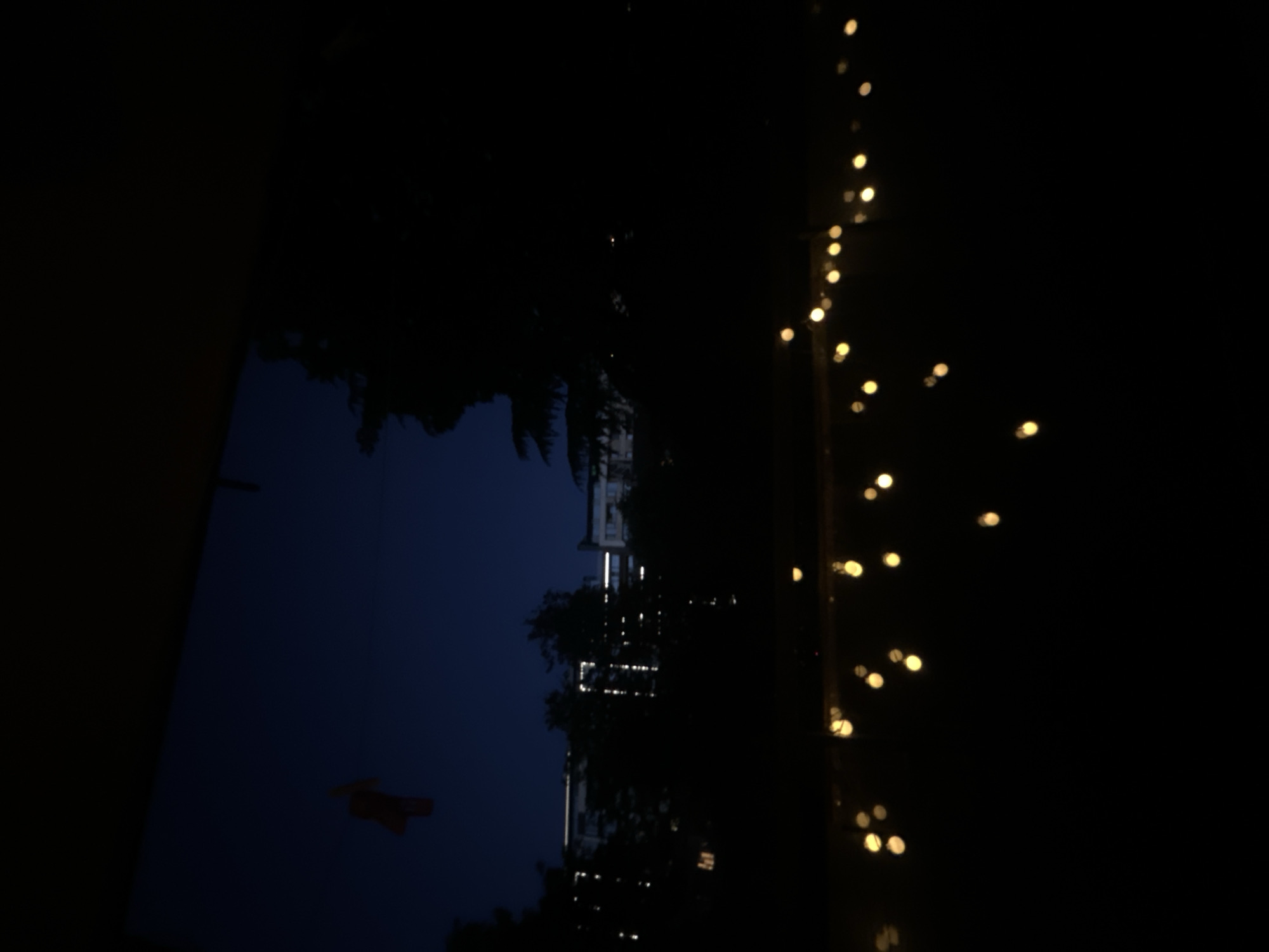 A nighttime view from a window shows a dark blue sky above the silhouettes of trees and buildings. In the upper left corner, a small, reddish-orange object, possibly a kite or decoration, hangs against the sky. In the middle ground, the illuminated windows of apartment buildings create bright, linear patterns against the dark structures. In the foreground, a string of yellow lights, blurred and out of focus, hangs low, casting a warm glow against the darkness. The scene appears to be an urban setting, potentially a residential area in Sofia, Bulgaria, as suggested by the architecture and the presence of trees and lights.