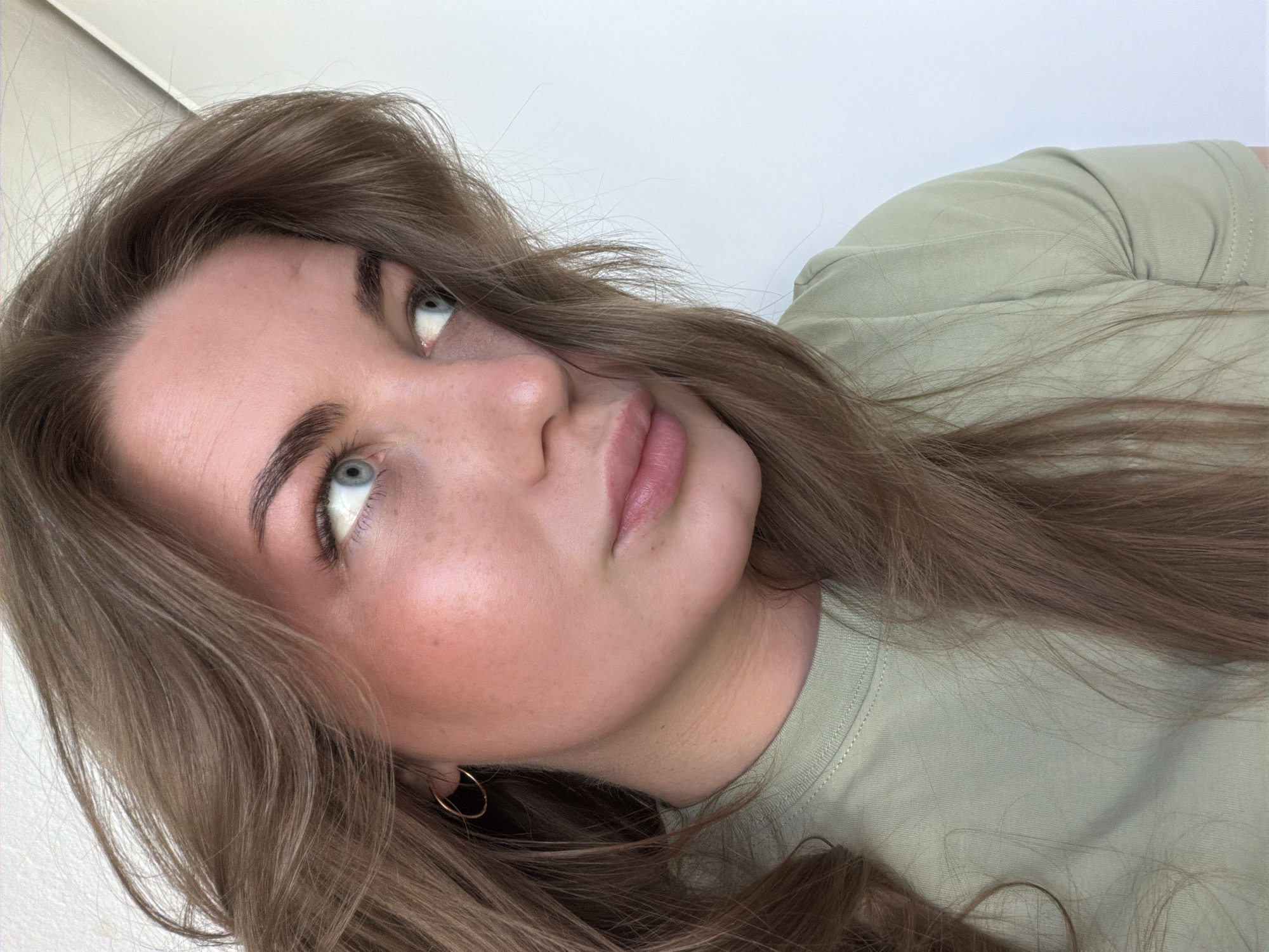 A young woman with long brown hair and light eyes is facing upward. She wears a pale green t-shirt and a small gold hoop earring in her left ear. Her skin is light with slight freckling on her cheeks. The background is a plain white wall. There is no evidence to suggest the location is in Chaikovsky, Russia.