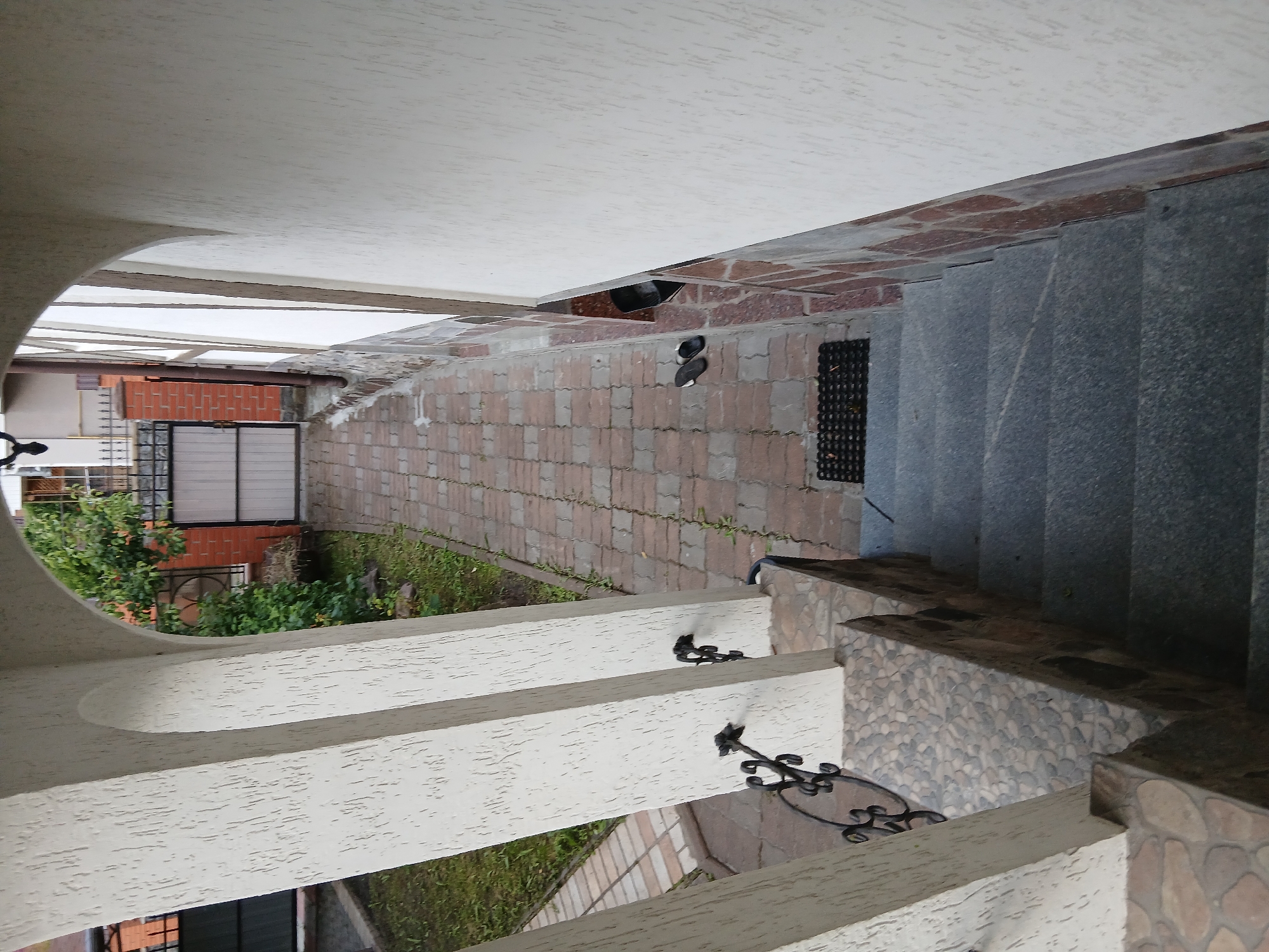 The image shows a view from an arched entryway looking down a paved path towards a gate. The entryway has thick, textured white columns. To the left of the path is a patch of green grass, and to the right is the textured wall of a building. The path is made of interlocking pavers and is lined with green grass. Further down the path, there is a metal gate with vertical bars and a solid metal panel, set between brick pillars. A tree with green leaves is visible behind the gate. There is a pair of dark-colored flip-flops on the paved path near a black doormat. The scene is set in Vita-Poshtova, Ukraine. The perspective suggests the photograph was taken from a slightly elevated position, possibly a porch or balcony.