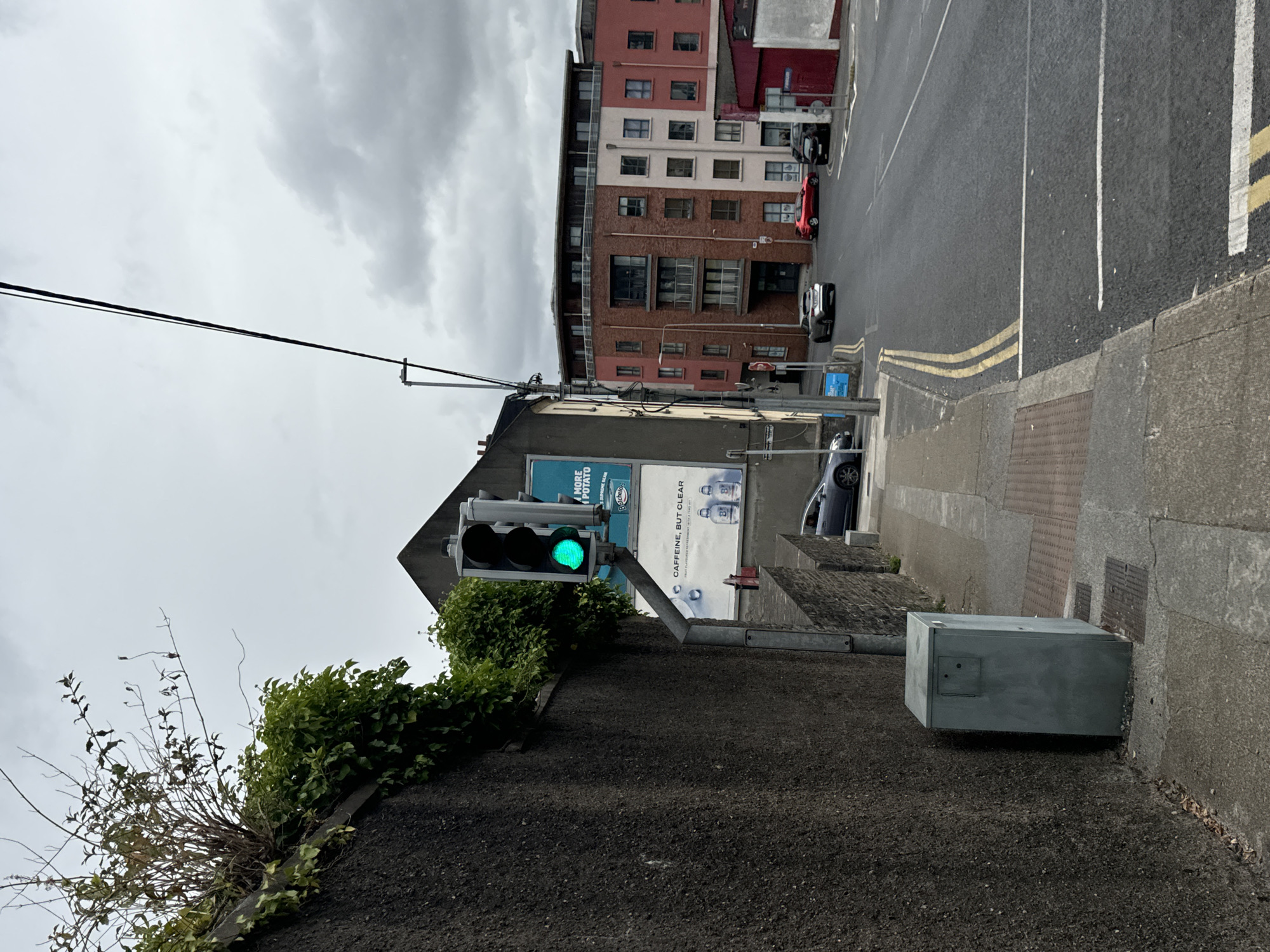 The image depicts a street scene in Waterford, Ireland. A traffic light showing a green signal is prominently positioned on the left side of the frame, attached to a pole extending from a concrete wall. Adjacent to the traffic light, a large billboard displays advertisements for "More Potato" and "Caffeine, But Clear." A grey electrical box is situated at the base of the wall. The street is paved with asphalt and marked with yellow and white lines, indicating lanes and a pedestrian crossing. Several cars are visible on the road, including a silver hatchback, a red car, and a black car. Buildings line the street, with a multi-story brick building on the right featuring numerous windows and balconies. The sky is overcast with gray clouds. The overall impression is a typical urban street view on a cloudy day.
