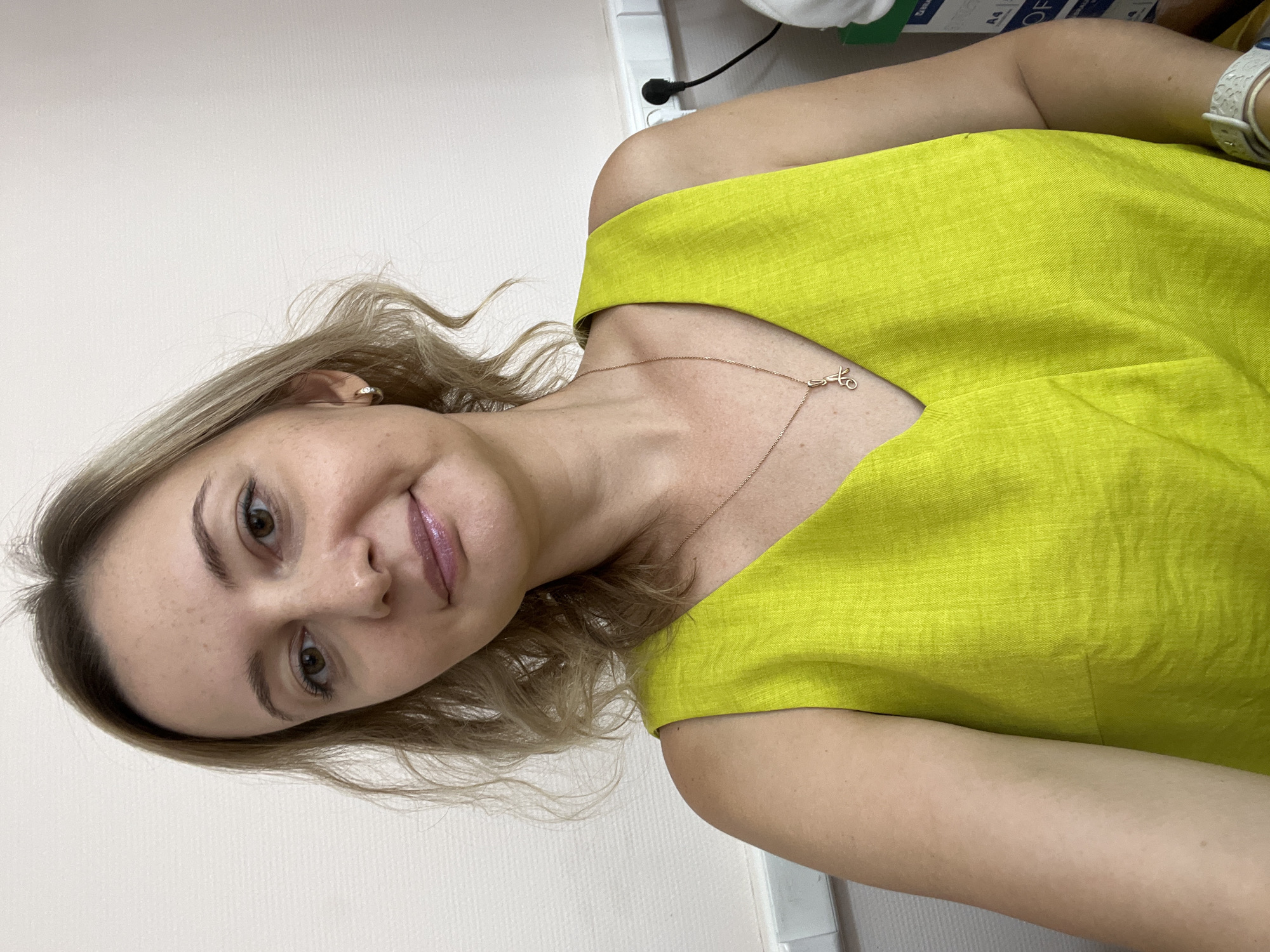 A woman with blonde hair and light brown eyes is pictured in a close-up selfie, wearing a sleeveless bright yellow dress. She has a light-colored necklace with a small pendant and a small hoop earring in her left ear. Her skin has some freckles. The background appears to be an indoor setting with a lightly textured off-white wall. There is a white horizontal strip near the bottom of the image and a black electrical cord with a plug extending from the wall. To the right, a portion of a white box with text is visible. The overall lighting suggests an indoor, daytime environment. The provided information about the image's location in Volgograd, Russia, cannot be visually verified from the image itself.