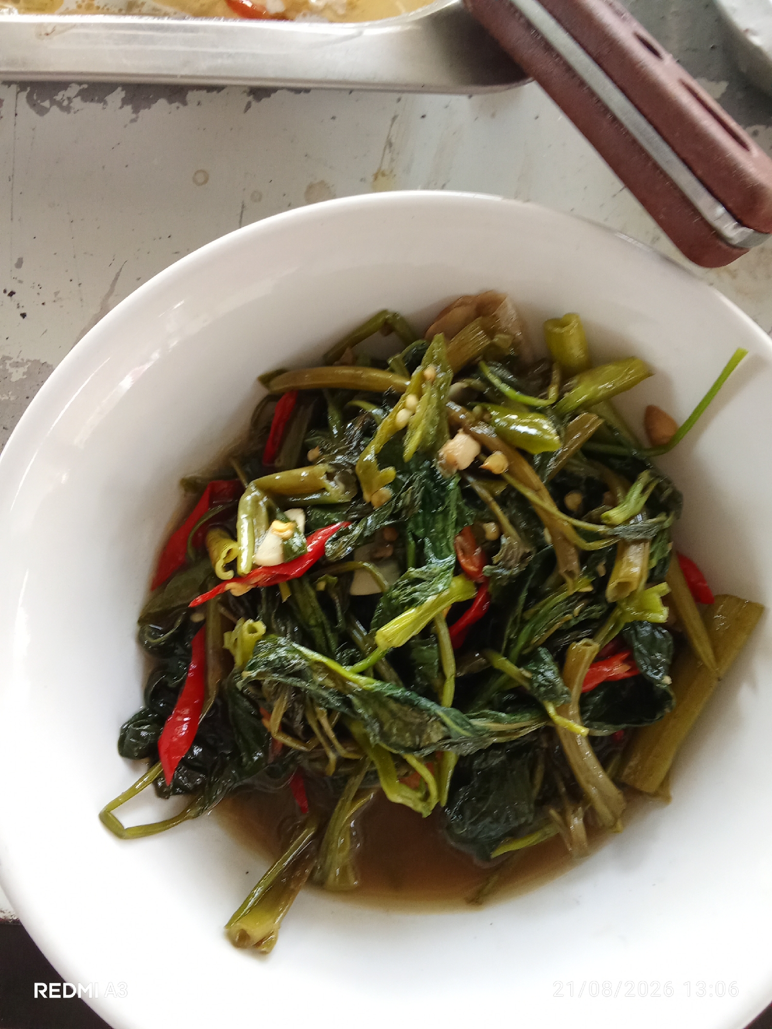 The image shows a close-up, overhead view of a white bowl filled with stir-fried water spinach (kangkung). The dish contains visible pieces of red chili and garlic. The bowl is placed on a textured, light-colored surface that appears to be a table with some stains and wear. In the background, a portion of a utensil with a brown handle and metal shaft is visible. The bottom left corner of the image displays the text "REDMI A3," and the bottom right corner shows the date and time "21/08/2026 13:06." The scene is lit from above, creating natural shadows within the bowl. No people are present in the image. The context of Banaran, Indonesia, is not directly evident from the visual information in the image itself, but the presence of water spinach and the likely Indonesian date format suggest a connection to Indonesian cuisine and location.