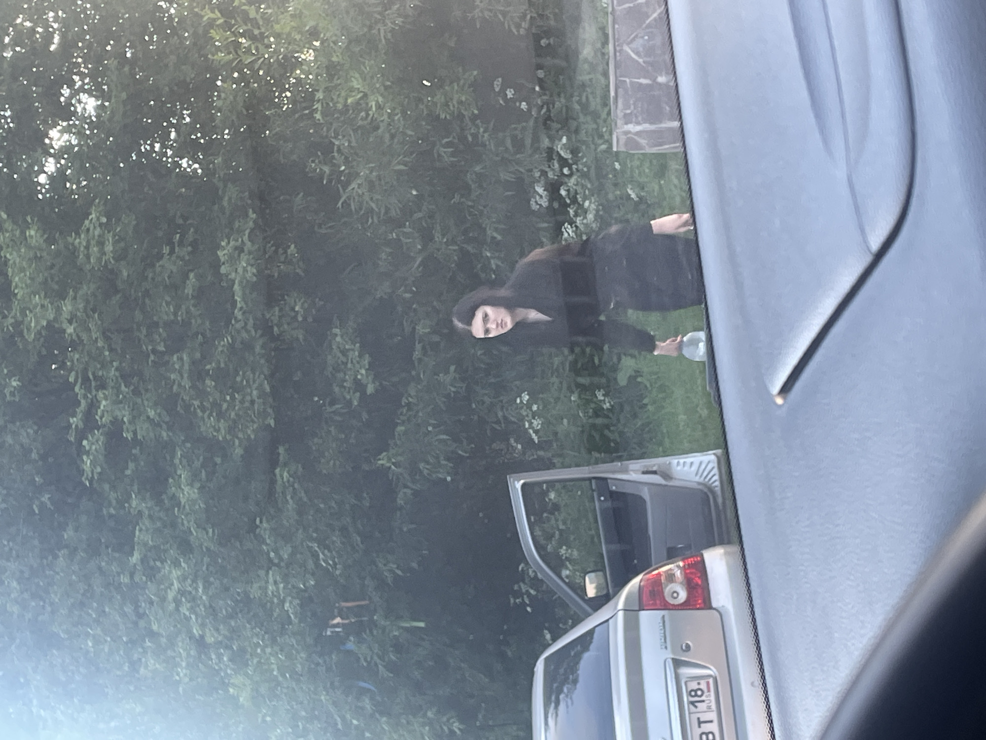 In this image, a person with dark hair, wearing a black hooded jacket, is seen bending down outdoors. The setting appears to be a grassy area with lush green trees and some white flowers in the background. The person is holding a clear, round object, possibly a glass or a bottle, in their hands.
In the foreground, the rear of a silver car is visible, with its driver's side door open. A license plate with Cyrillic lettering and numbers, reading "BT 18. RUS", is attached to the back of the car. The overall lighting suggests it is daytime, possibly late afternoon or early evening, given the soft light filtering through the trees. The image is taken from inside another vehicle, as indicated by the reflection on the window. The scene is set in Gavrilovka, Russia, suggesting a rural or suburban environment. There are no clear indications of the weather conditions, but the greenery suggests it is not winter. The person's posture and the object they are holding do not convey any specific emotion.
