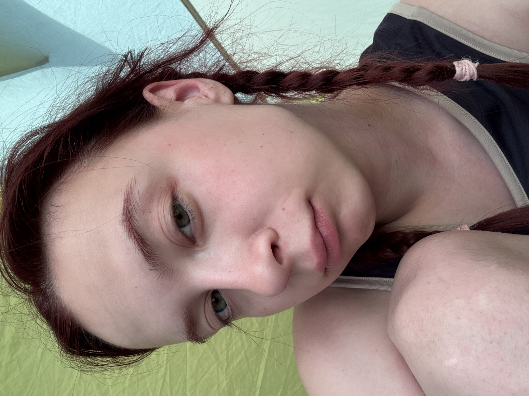 A fair-skinned person with auburn hair, styled in two braids secured with light pink ties, is indoors, possibly in a tent given the green and blue background. The person is wearing a dark tank top with a light-colored trim. The viewpoint is a close-up, slightly tilted angle. No specific information connects this image to Khvalynsk, Russia.