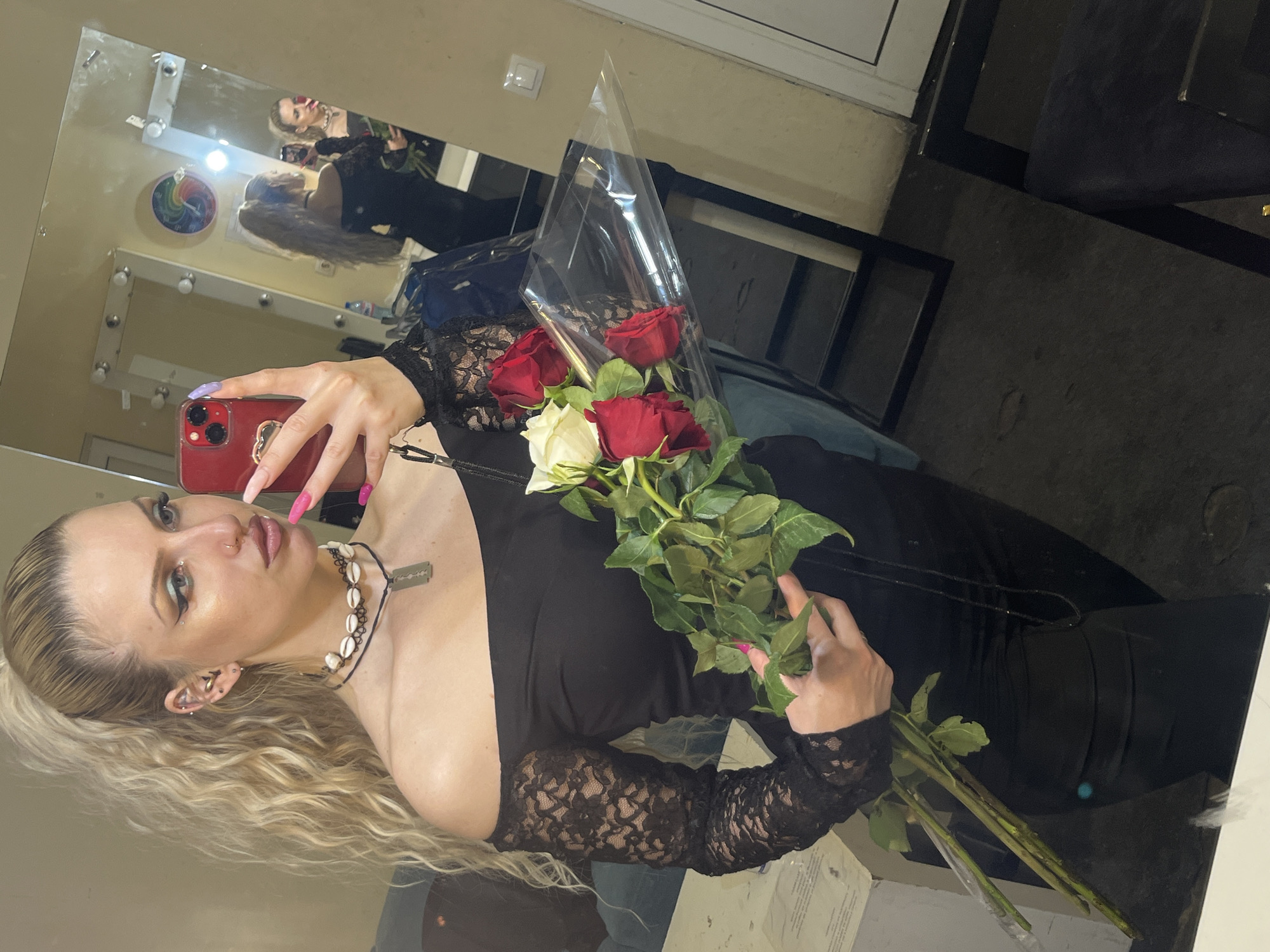 The image depicts a woman holding a bouquet of roses in what appears to be a dressing room. She is taking a selfie with a red phone, capturing her reflection in a makeup mirror. The woman has blonde hair pulled back in a ponytail and is wearing a black off-the-shoulder dress with lace sleeves. A shell necklace with a razor blade pendant adorns her neck. The bouquet consists of red and white roses partially wrapped in clear plastic.
The reflection in the mirror shows the woman and the bouquet again, along with a glimpse of a colorful circular object on the wall near the mirror. The room has neutral-colored walls, and the mirror is surrounded by light fixtures. The environment is somewhat cluttered, with glimpses of furniture and items in the background. The location is not evident based on the image content. The request for information on the city of Plovdiv, Bulgaria, is irrelevant as there is no evidence of the city.
