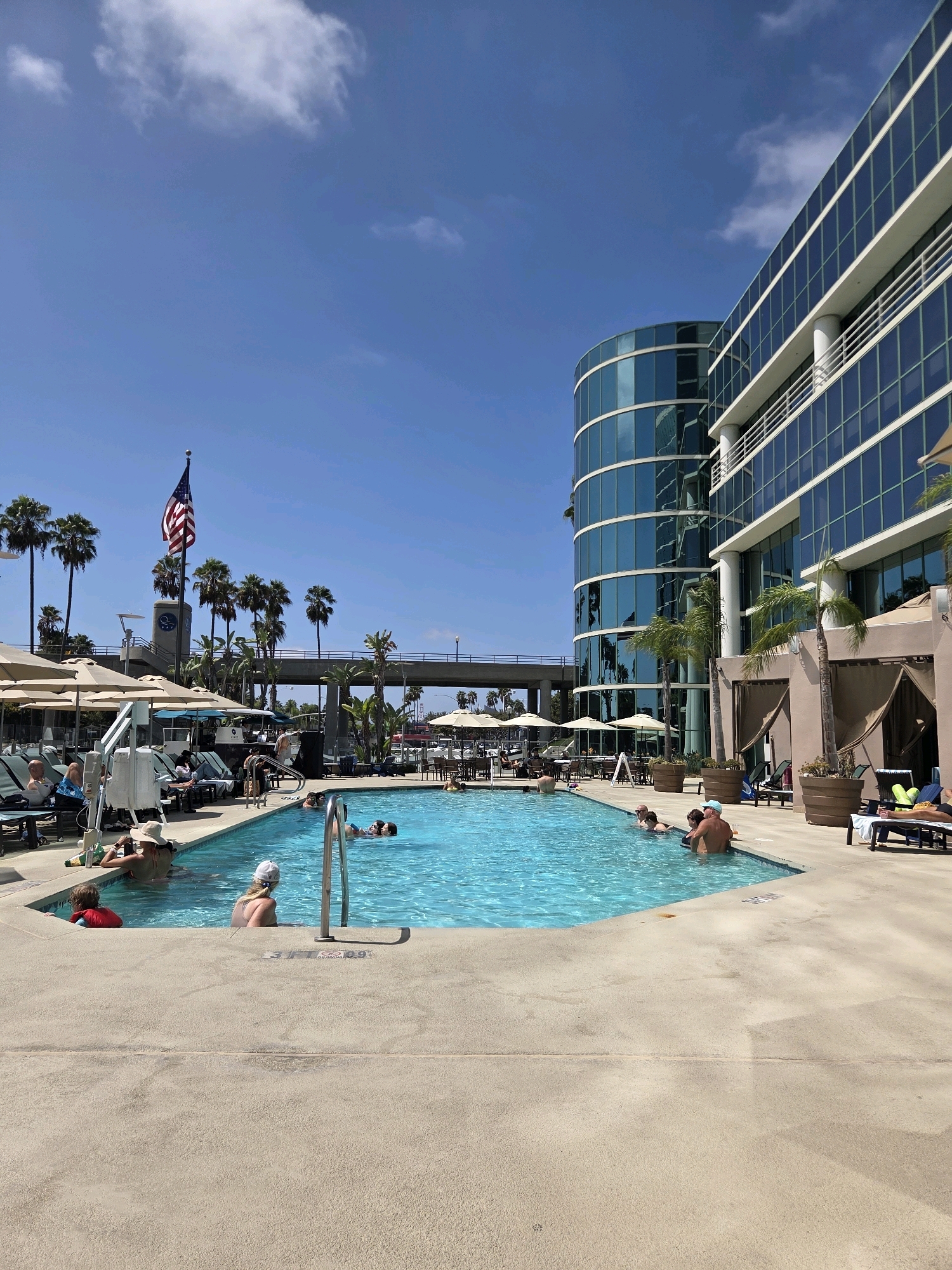 The image shows a sunny day at a hotel pool in Long Beach, California. In the foreground, a large, bright blue swimming pool is populated by several people. Some are standing in the shallow end, while others are swimming or relaxing in the water. There are lounge chairs and umbrellas arranged around the pool area, with some people seated or lying on the chairs, enjoying the weather.
In the background, a tall, modern building with a curved glass facade dominates the skyline. The building's blue-tinted windows reflect the clear sky. An American flag is prominently displayed on a flagpole near the pool, adding a patriotic touch to the scene. Palm trees are scattered throughout the area, typical of the Southern California landscape. An elevated walkway or bridge is visible in the distance, suggesting the proximity of transportation infrastructure. The overall atmosphere is one of relaxation and leisure, with people enjoying a warm day outdoors.