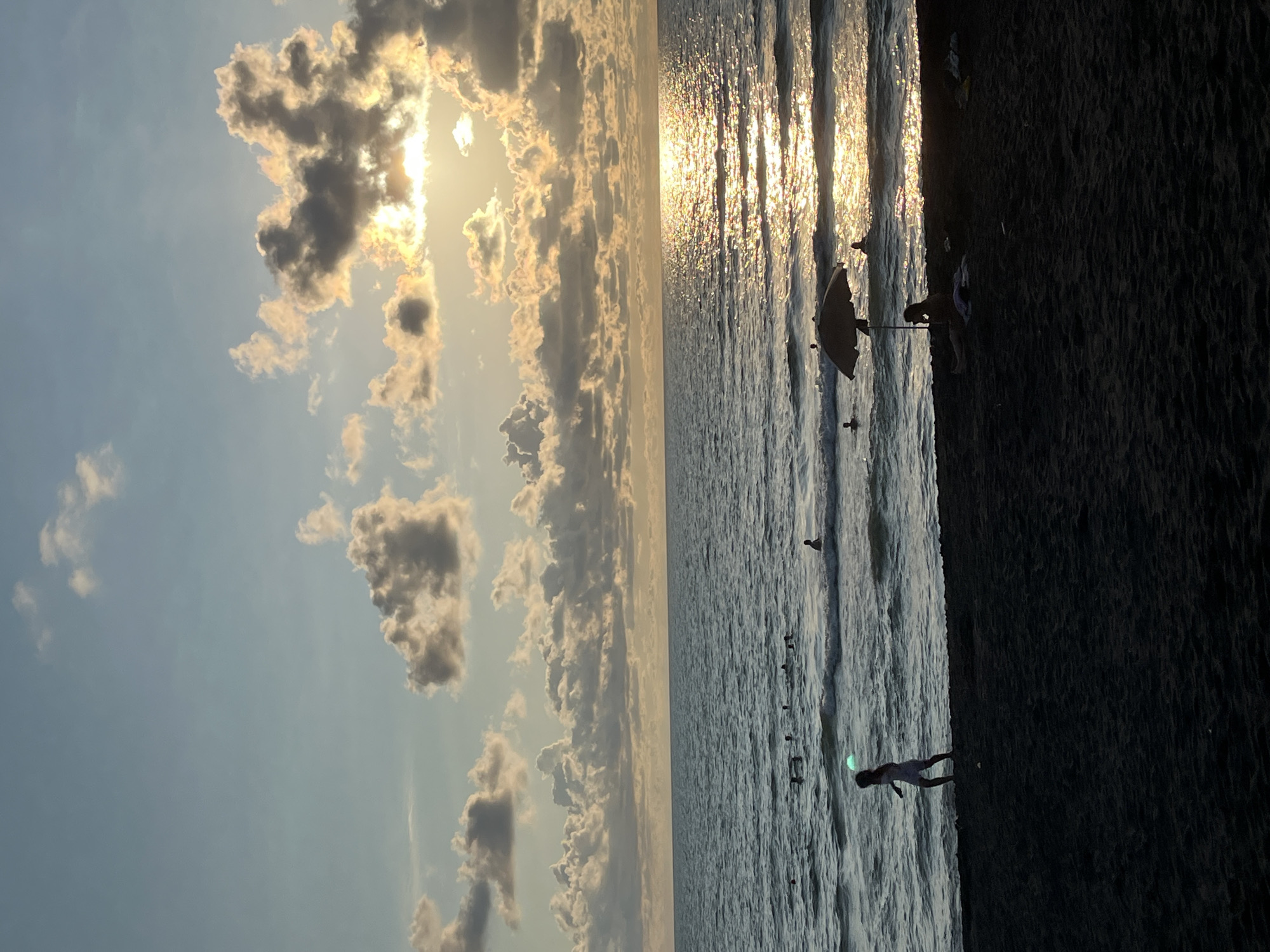 The image depicts a beach scene in Ureki, Georgia, during what appears to be sunset. The foreground is a dark, sandy beach. To the left, a child in a light-colored dress is running along the shoreline. In the middle ground, several people are visible in the ocean, swimming and wading. An umbrella is set up on the beach with a person sitting beneath it. The ocean is a deep blue with moderate waves, and sunlight reflects off the water, creating a shimmering effect. The sky is a pale blue with scattered, fluffy clouds illuminated by the setting sun, which is partially obscured by clouds, casting a warm, golden glow. The overall atmosphere is serene and recreational.