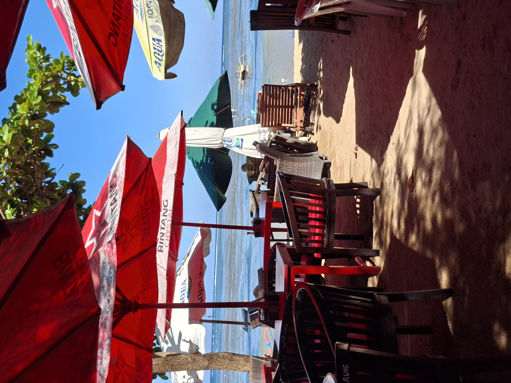 This photograph captures a sun-drenched beach scene, likely in Denpasar, Indonesia. The perspective is from under the shade of numerous red and white umbrellas advertising "Bintang Crystal" and "Aqua," suggesting a beachside establishment. The foreground is filled with the dark wooden frames of chairs and tables, creating a sense of depth as the eye moves towards the beach. The sand is light tan, marked by shadows from the umbrellas and furniture. In the distance, the blue water of the ocean is visible, with a traditional outrigger boat anchored near the shore. The bright blue sky above contrasts with the red umbrellas, and the clear water indicates a sunny day, creating a tranquil atmosphere of a tropical beach resort.