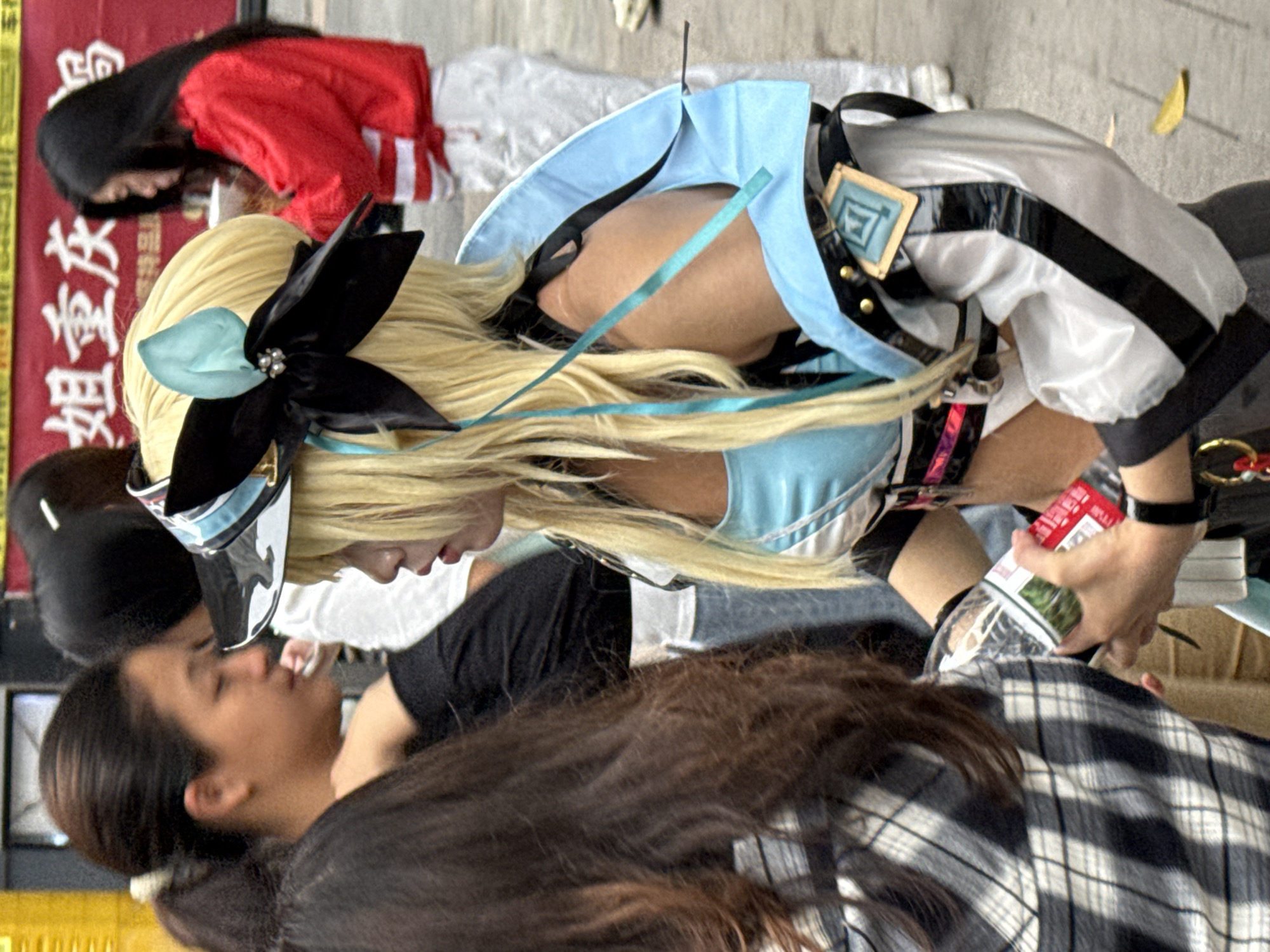 The image captures an outdoor scene in what appears to be the city of Yuzhong District, China. The photo primarily features a person with long, blonde hair wearing a costume, possibly for cosplay. The costume includes a light blue top, a black and white jacket with a square emblem on the shoulder, and a large black bow with a light blue accent in the hair. The person is bending over or leaning forward.
Several other people are visible in the background and foreground. A person with long, dark hair wearing a red jacket is partially visible behind the costumed person. In the foreground, a person with long, dark hair is wearing a black and white plaid shirt.
The background includes what appears to be a sign with Chinese characters, indicating the location in China. A clear plastic bag containing some kind of packaged food or product is visible near the hand of the person in the costume. The overall environment appears to be a public area, possibly a street or gathering space.