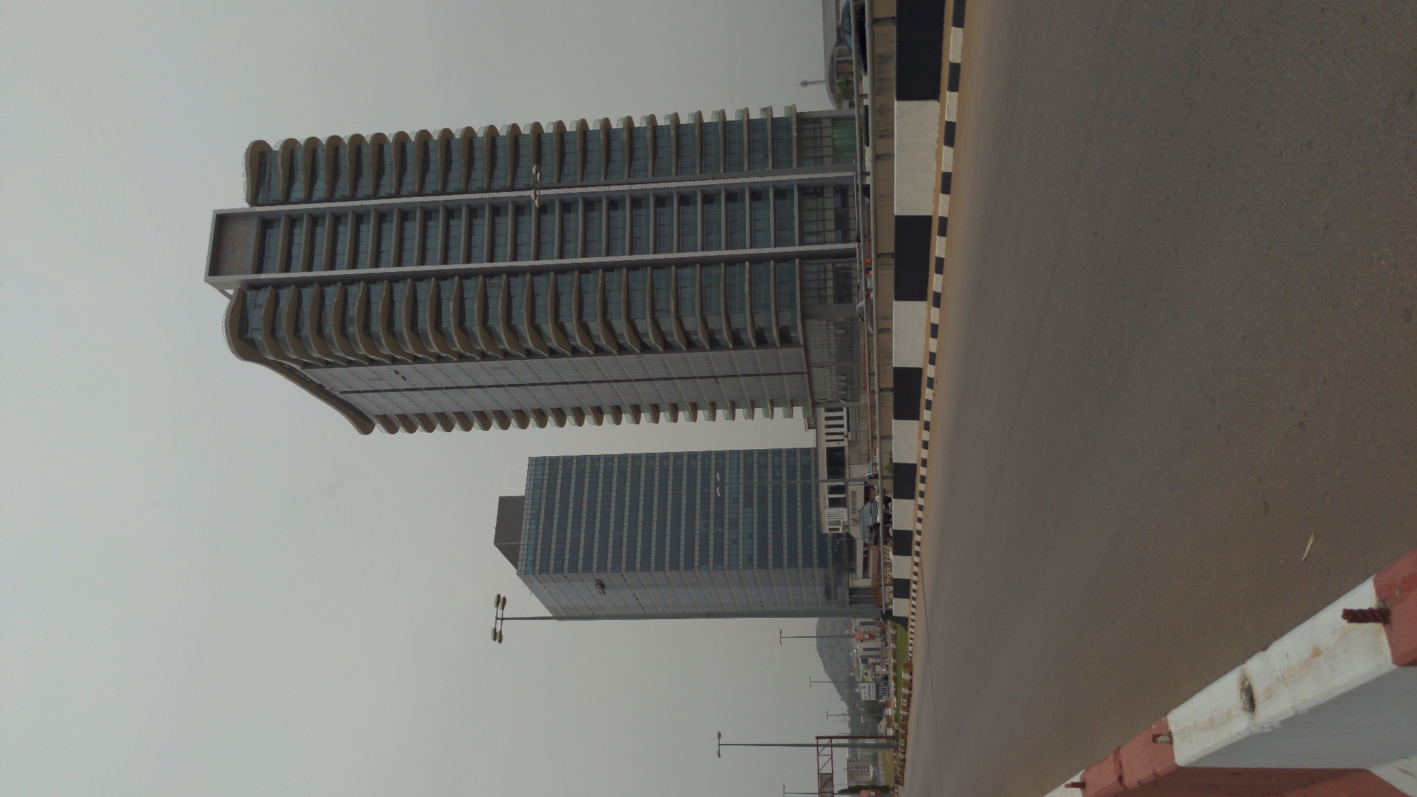 The image depicts a modern urban landscape in Abuja, Nigeria, under an overcast or hazy sky. A wide, multi-lane asphalt road spans the right side of the frame, with a black and white striped curb marking its left edge. A red and white concrete barrier is visible along the far right foreground of the road.
Two prominent high-rise buildings stand on the left side. The building closer to the viewer features a distinctive modern architectural design, characterized by multiple horizontal levels and a facade adorned with numerous vertical, curved, fin-like structures. These elements give the building a unique, dynamic appearance. Behind it, a second, taller and more slender building is visible, featuring a more conventional grid pattern of windows. A faint human figure appears to be working on the facade of this rear building, possibly performing maintenance or cleaning.
Several contemporary streetlights with double arms are spaced along the road, extending into the distance. A few indistinct human figures can be seen at the base of the buildings, suggesting pedestrian presence. The sky is uniformly gray, indicating a cloudy or hazy day, which casts a soft, diffused light over the entire scene. There is no discernible text visible on any of the buildings, signs, or other objects in the image. The scene is calm and depicts a typical daytime urban environment.