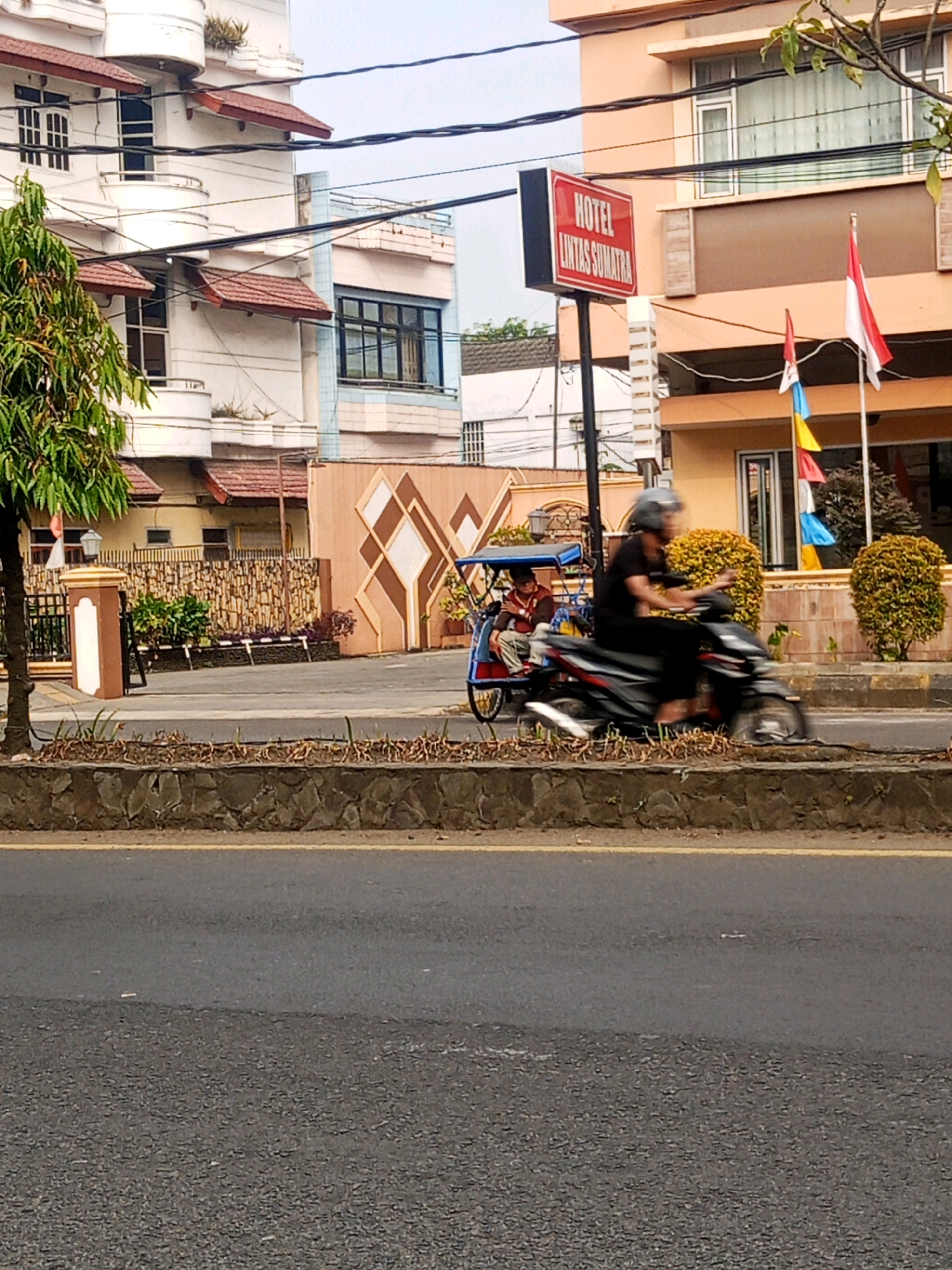 The image depicts a street scene in Lubuklinggau, Indonesia. In the foreground, a street with a yellow lane marker is visible, bordering a stone-lined embankment. Behind the embankment, a motorcycle with a blurry rider is in motion, accompanied by a three-wheeled vehicle, possibly a becak, with a seated passenger. The background reveals a building with a prominent red sign that reads "HOTEL LINTAS SUMATRA." This hotel building has a peach-colored facade with windows and balconies. Several Indonesian flags are displayed on flagpoles in front of the hotel. The scene also includes a tree with green foliage to the left, and decorative fencing and a wall with a diamond pattern further back. The overall environment suggests a commercial area in a city.