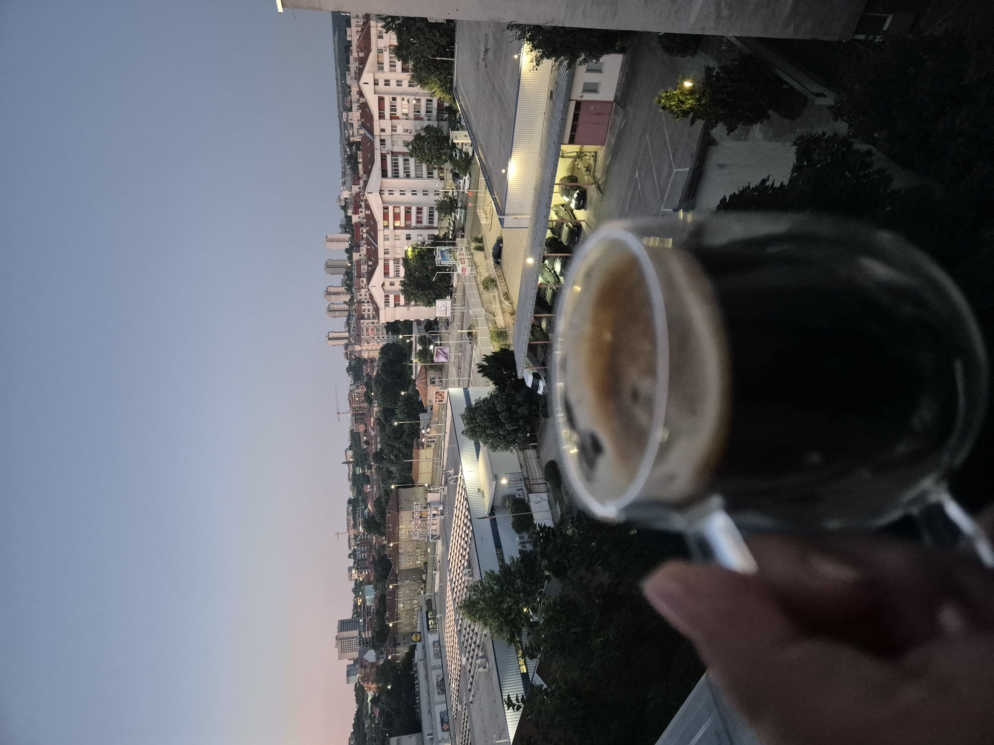 An outdoor, eye-level, medium shot shows a hand holding a double-walled glass mug of dark liquid, likely coffee, in the foreground, slightly out of focus. The mug has a clear handle on the right side. The background is an urban landscape at dusk. A flat-roofed commercial building with a large, lit facade and a sign that reads "LIDL" is prominent in the mid-ground. Adjacent to it is another commercial building with white siding. Beyond these, a cluster of residential apartment buildings with red-tiled roofs and white facades are visible. In the far distance, several taller buildings and cranes suggest a developing urban area. Streetlights are on, casting a warm glow on the scene. The sky is a gradient of pale blue and pink, indicating twilight. Trees are scattered throughout the landscape. A parked white van is visible near the commercial buildings, and cars are parked in lots.
