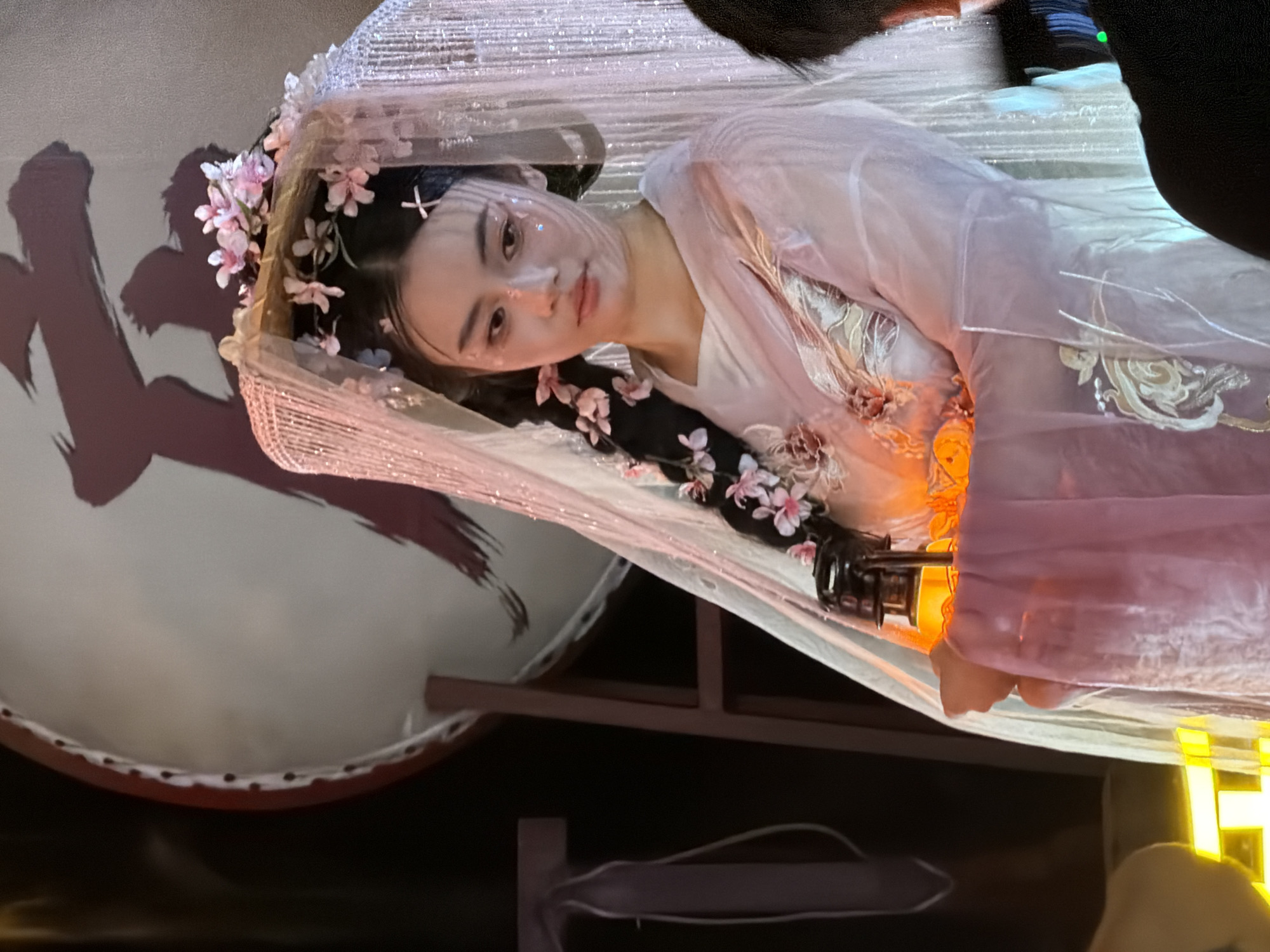 A young Asian woman with dark hair styled into braids adorned with pink flowers is looking down and to her right. She is wearing a light purple, sheer robe with intricate embroidery on the sleeves and front. A translucent veil, also decorated with small pink flowers, covers her head. In her hands, she holds a small, antique-looking lantern that emits a warm, orange glow. Her face is delicately made up with glitter accents on her cheeks and forehead. Behind her, a large, circular object, possibly a drum or a screen, features a dark, calligraphic brushstroke. The setting appears to be indoors, possibly a performance space or a themed event, with hints of stage lighting and dark backgrounds.