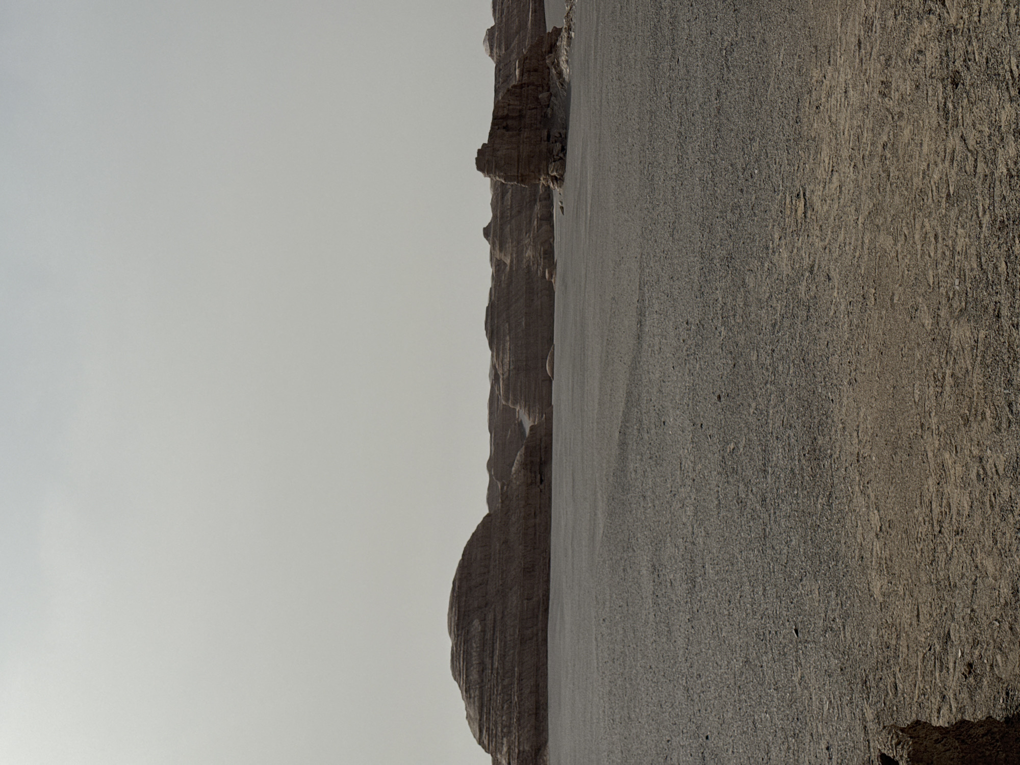 The image depicts a desert landscape in Dunhuang City, China. The foreground is dominated by a vast expanse of gravelly, sandy ground. To the left of the frame, a series of eroded rock formations rise against a pale, overcast sky. The formations vary in size and shape, with some resembling rounded hills and others appearing as jagged, layered cliffs. The scene is devoid of people or any discernible human presence. The overall impression is one of a remote, arid, and weathered environment.