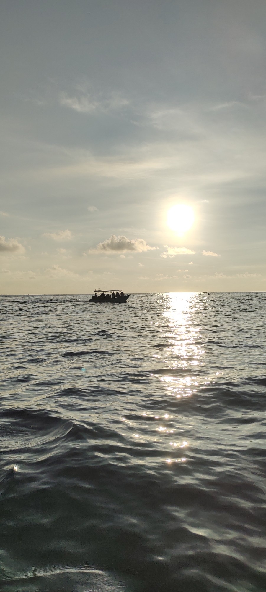 A wide shot captures a body of water with gentle waves reflecting the bright sun on the horizon. A small boat, silhouetted against the sun, carries several passengers and is positioned on the water. The sky above is a soft gray with scattered clouds. In the distance, another small object, possibly another boat or marker, is visible on the water. The overall scene suggests a serene moment at sea, likely near the city of Sabang, Indonesia, given the tropical lighting and coastal environment.