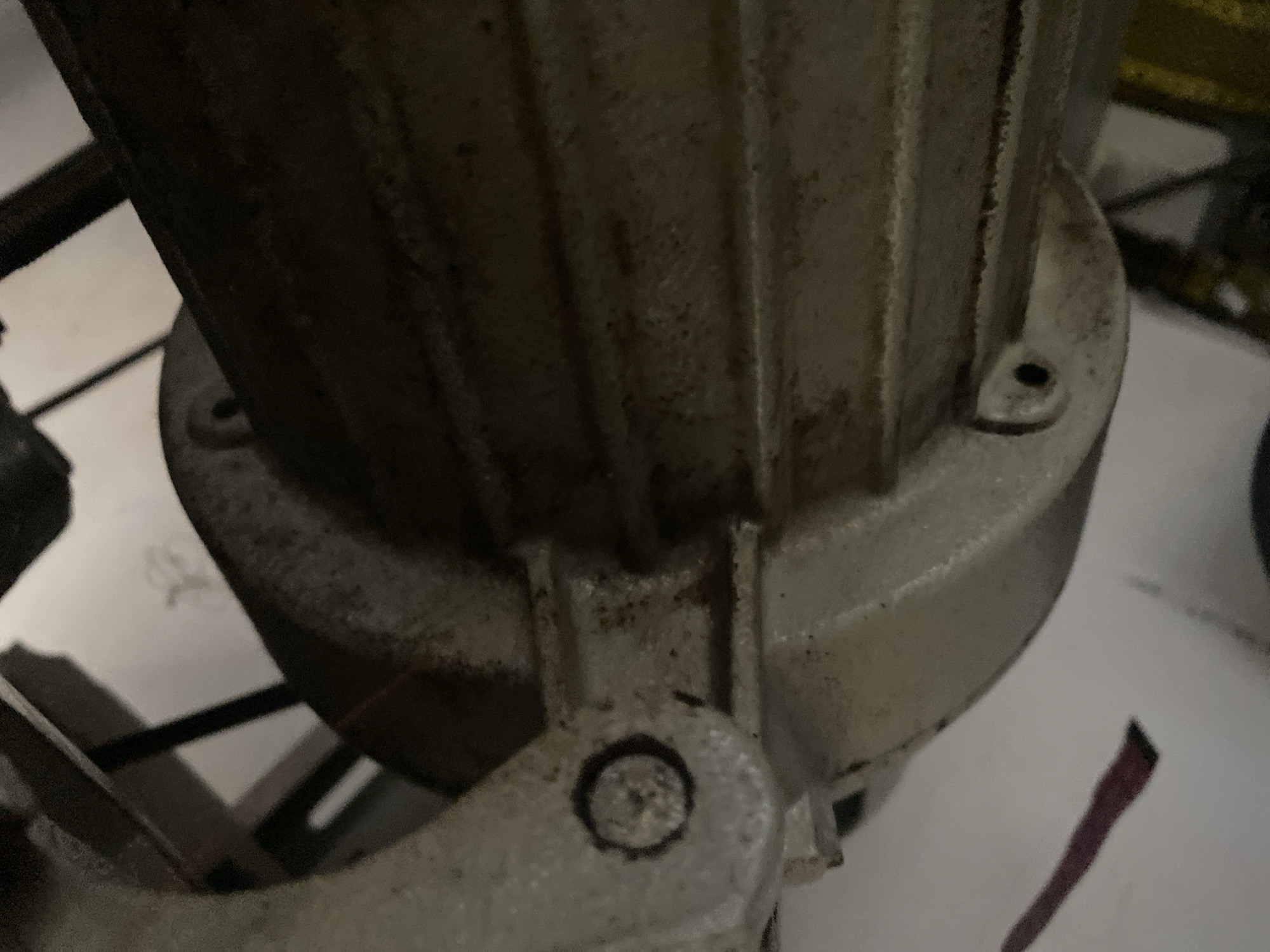 The media file shows a close-up, low-angle shot of a weathered, metallic object, possibly a motor or pump housing, with visible signs of age and use. The object is predominantly silver-grey and exhibits a ribbed texture along its cylindrical body. There are dark stains and dirt accumulated in the grooves and crevices, indicating it has been exposed to environmental elements or is part of a working mechanism. The base of the object is wider and appears to be attached to a mounting bracket with a visible screw. Another screw is present on a protruding lip on the side. The background is mostly out of focus, revealing indistinct shapes and colors, including white surfaces and dark, possibly yellow, structures in the upper right. The lighting is dim, creating shadows that emphasize the texture and wear of the object. The overall impression is of an industrial or mechanical component in a somewhat neglected state. No people, specific activities, or discernible text are visible in the frame. The location cues suggest an industrial or workshop setting in Jalingo, Nigeria, but this is not definitively confirmed by visual elements within the image.