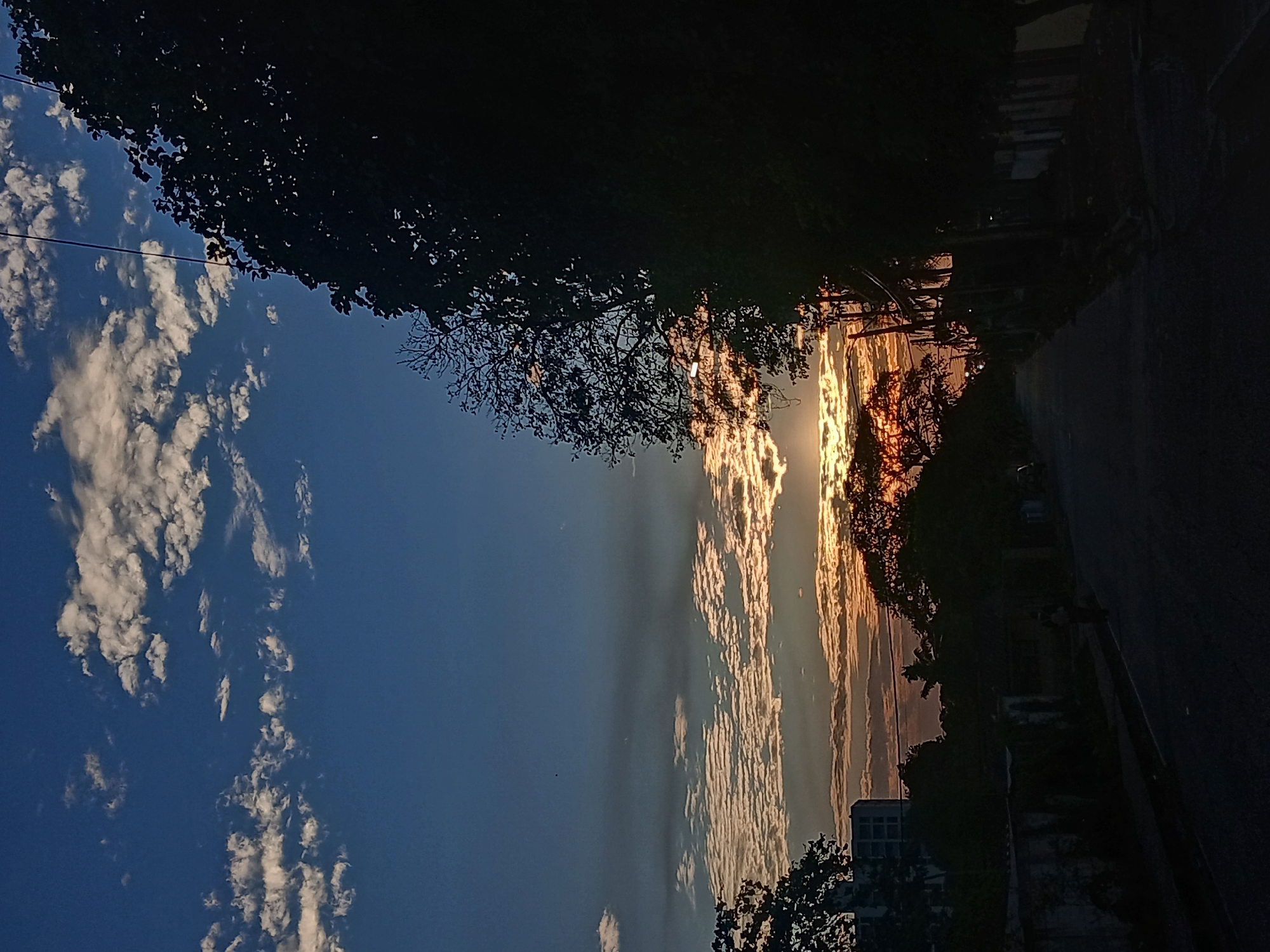 The image depicts a sunset scene along a street in what is presumed to be Juan Griego, Venezuela. The sky is a gradient of blue to orange and gold, filled with elongated, striated clouds illuminated by the setting sun. Silhouetted trees and buildings line the right side of the image, defining the edge of the street. A road runs along the right side, leading to a dim street and some people walking near the sidewalk. A power line crosses the sky on the left. The lighting suggests the photograph was taken during either dawn or dusk.