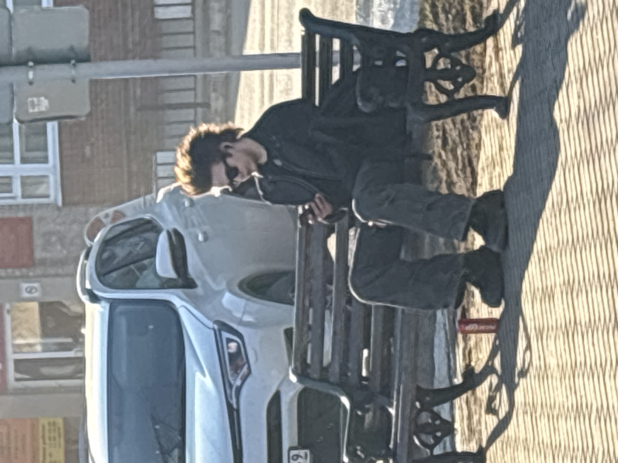 A man reclines on a black bench in front of a white car. The bench is cast iron and wood. The man wears a black jacket and dark pants. The scene is located in the city of Chaikovsky, Russia. The background shows a building and a traffic light pole. The ground is paved with a patterned surface. A red object is near the edge of the bench. The car appears to be parked on the street.