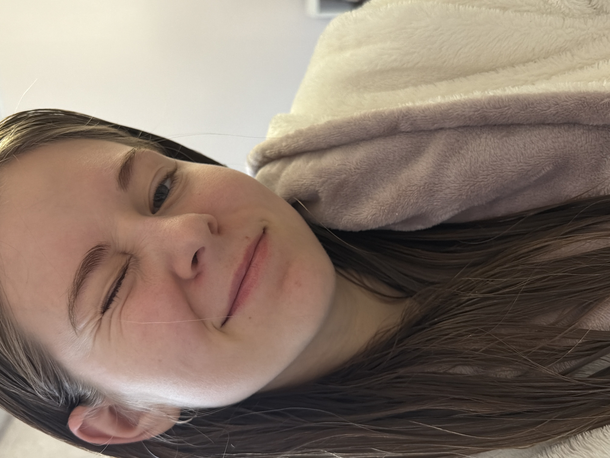 A playful close-up portrait shows a young person, likely female, with fair skin and long, straight, medium brown hair, rotated 90 degrees counter-clockwise. The individual is winking with their left eye, creating noticeable crinkles around it, and smiling subtly. Their right eye is open and looks towards the viewer. Several fine strands of hair are visible across their face and extending from their hair.
They are enveloped in a soft, plush fabric, likely a blanket or robe, which has a cream-white upper section and a light brown or taupe lower section. The background is a stark, bright white, suggesting an indoor setting with soft, ambient lighting, characteristic of daylight.
The dominant emotion conveyed is one of happiness and mischief. No text is visible in the image. The geographical context for this image is Perm, Russia.