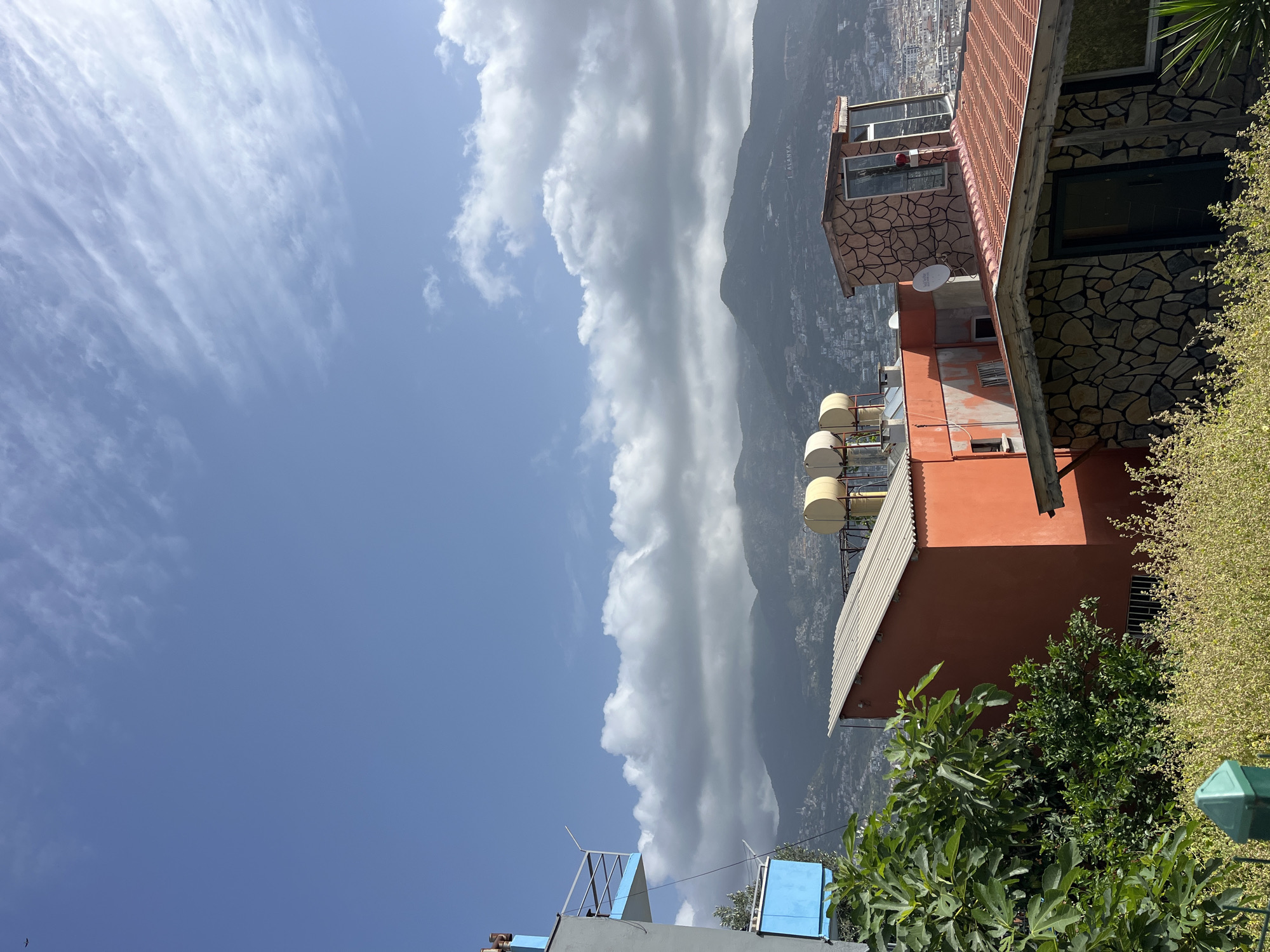 The image depicts a view in Alanya, Turkey, focusing on buildings and the landscape. In the foreground, there are buildings with various architectural elements, including stone facades, red roofs, and solar water heaters. Lush greenery and trees partially obscure the lower sections of these buildings. In the background, a mountain range is visible, and the skyline is partially obscured by a thick line of white and grey clouds. The sky above is a mix of clear blue and wispy clouds. The overall impression is that the photo was taken during the day, with a view overlooking the city.