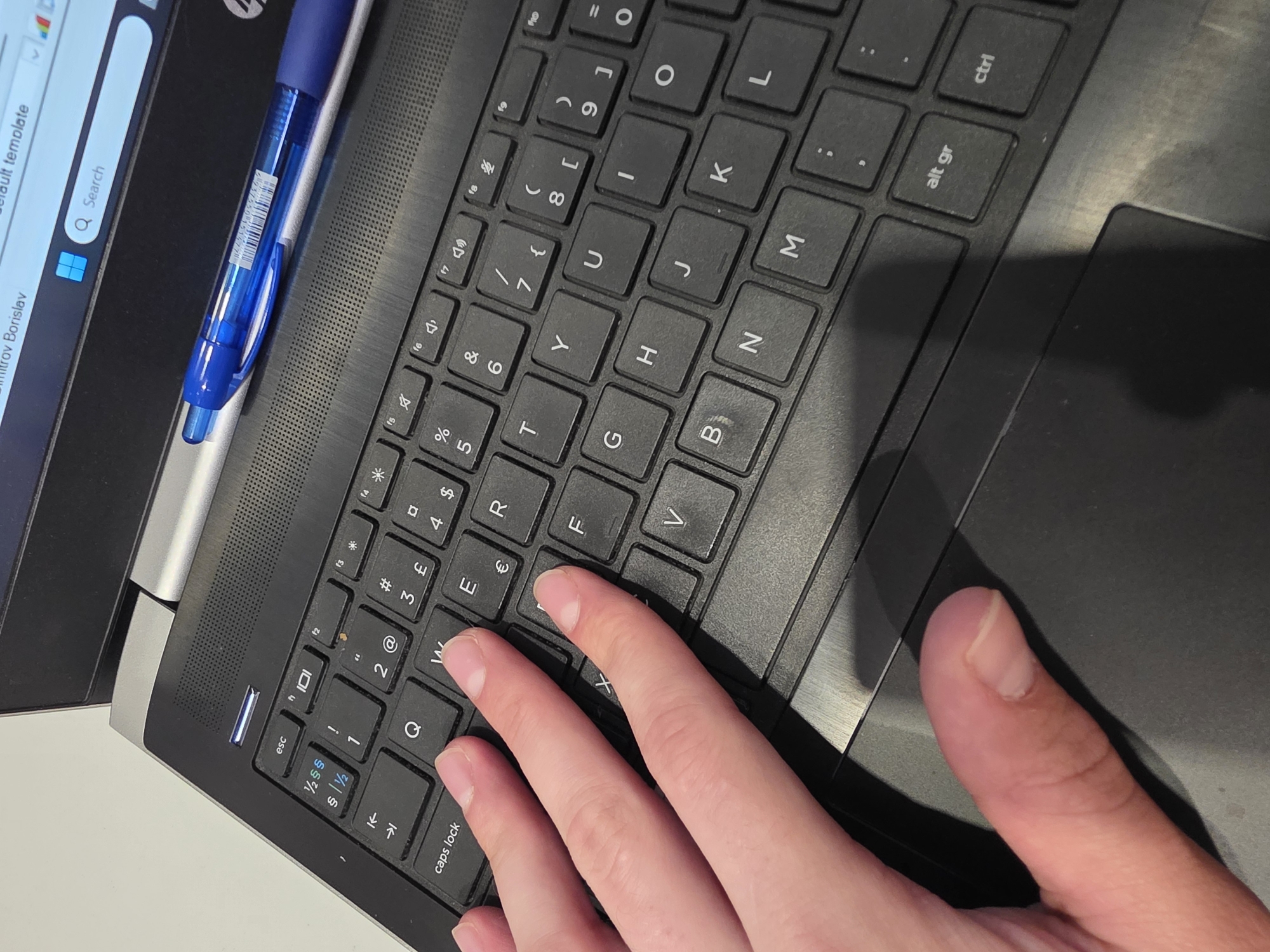 The image shows a person's hand typing on a laptop keyboard. The laptop is silver and black and has a blue pen lying on the left side. The person's fingers are pressing the "W", "E", and "F" keys. The trackpad is visible on the right side of the keyboard. A window is open on the screen, displaying a search bar and the name "Dimitrov Borislav". The location is not determinable from the image.