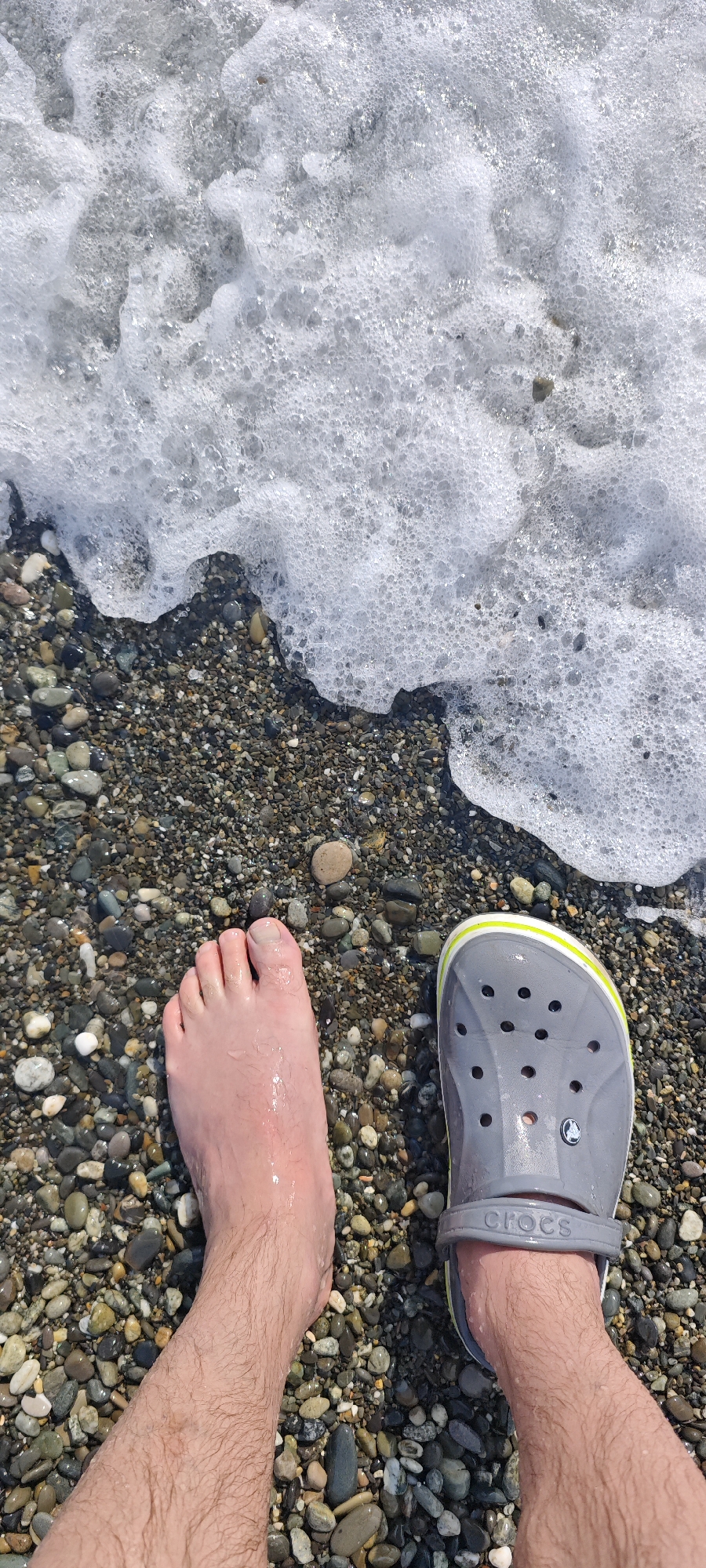 The image shows a first-person perspective of a person standing on a pebble beach with the surf washing around their feet. One foot is bare, wet with seawater, while the other wears a grey Crocs shoe with a light green and yellow trim. The beach consists of various-sized pebbles in shades of grey, brown, and tan. The surf is foamy and white. The location is likely Sochi, Russia, given the Black Sea's pebble beaches characteristic of that region. No other people or specific landmarks are visible.